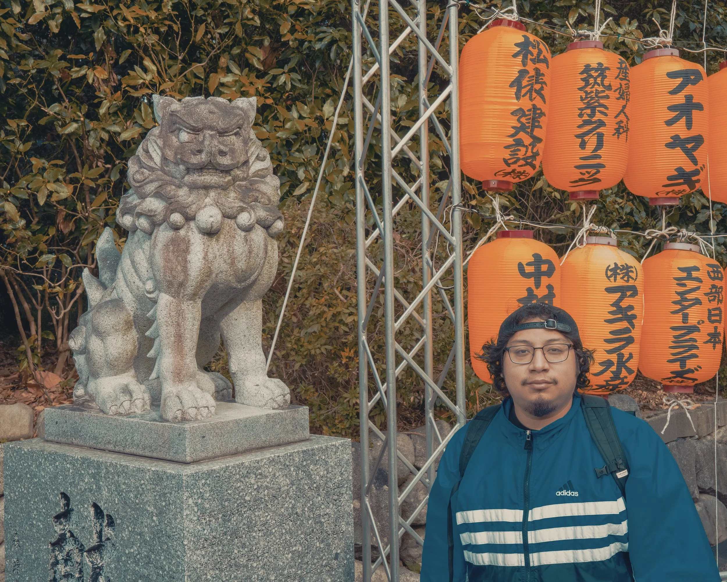 A man with glasses, a black cap, and a blue Adidas jacket stands in front of a stone statue of a lion and orange Japanese lanterns with black kanji writing, surrounded by greenery.