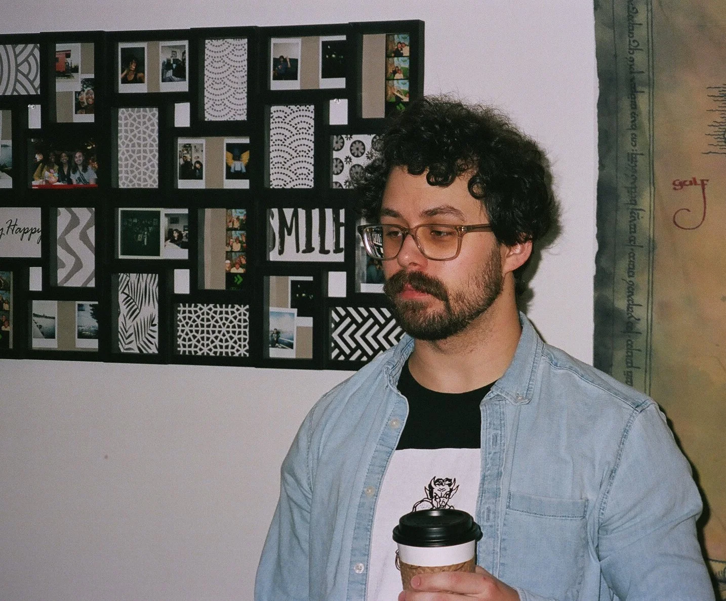 A man with glasses and curly hair holding a coffee cup, standing in front of a wall with a photo collage and decorative patterns.
