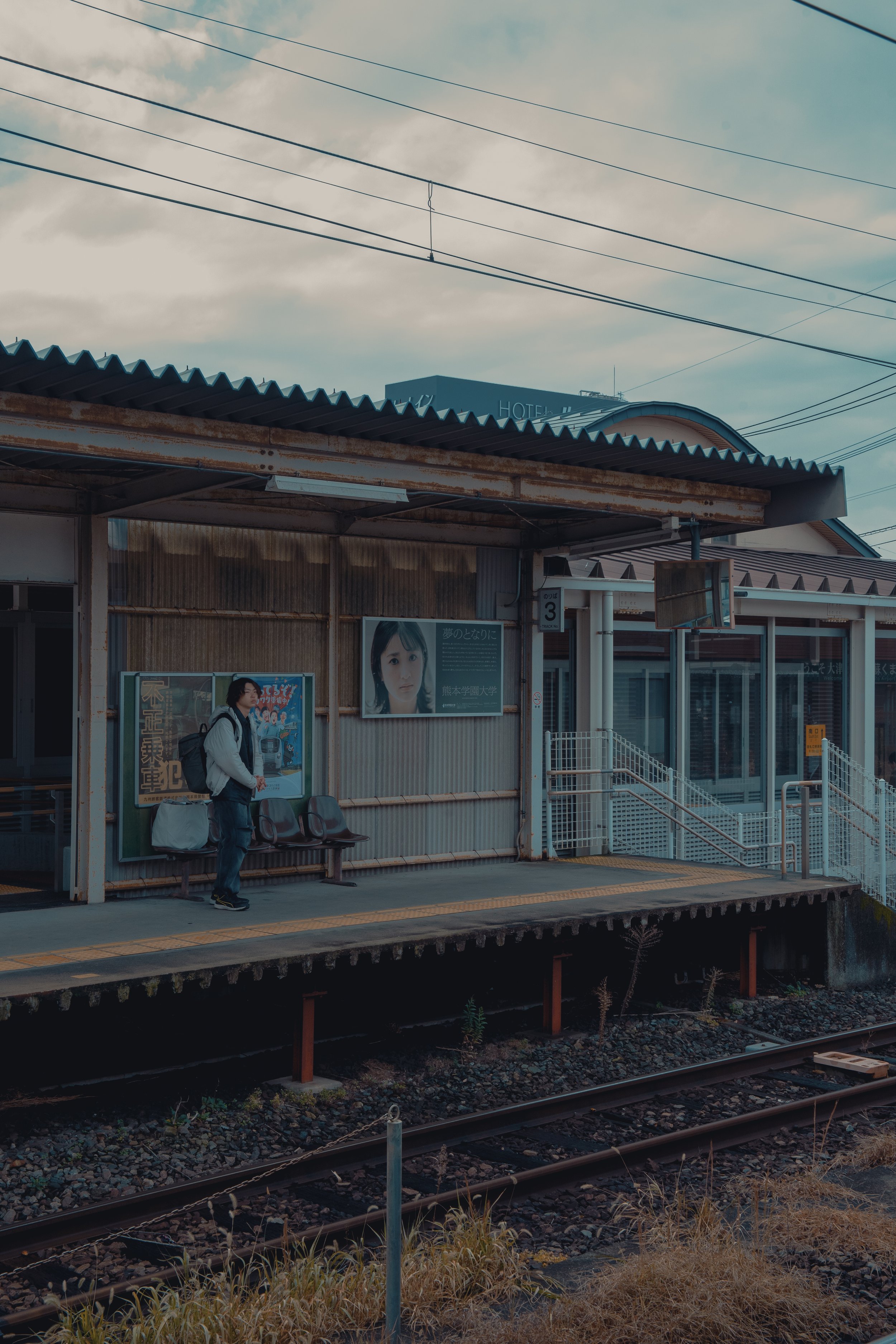 A woman standing on a train station platform with a backpack, next to a wall with posters, overlooking train tracks underneath a rusty roof, on a cloudy day.