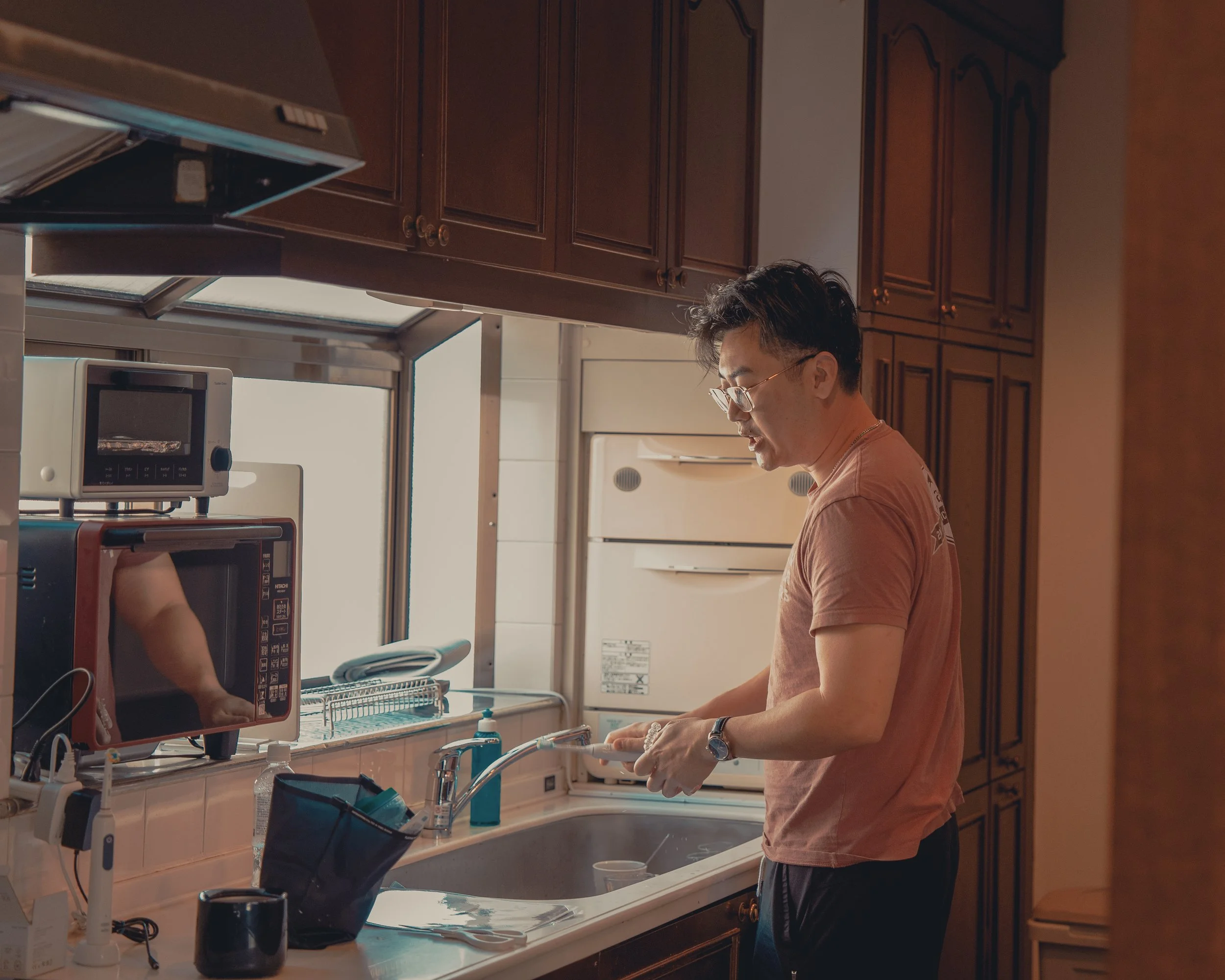 A man with glasses in a kitchen washing dishes at the sink, with a microwave and other appliances nearby.