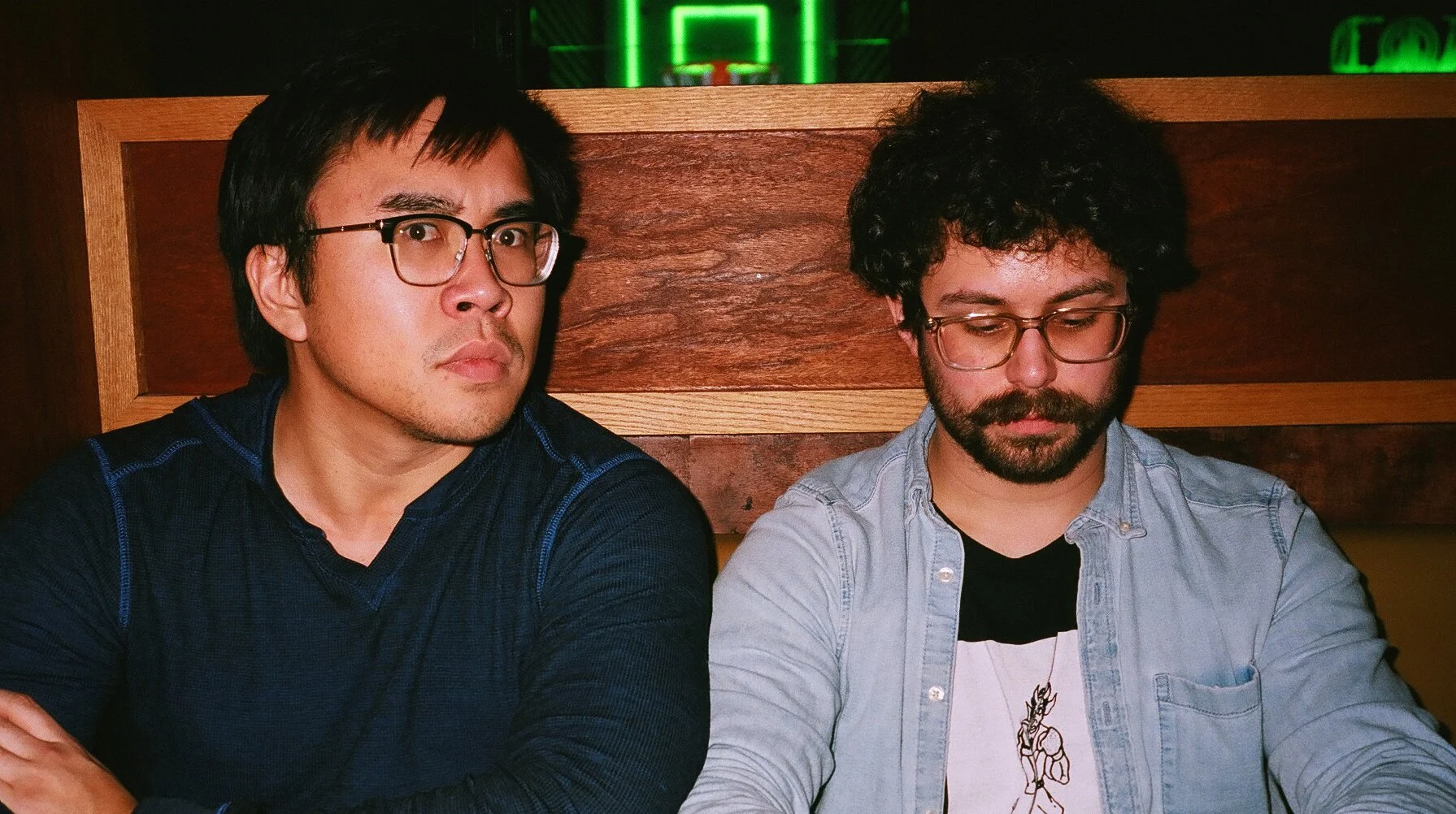 Two men sitting at a table in a dimly lit restaurant or bar, with wooden panels behind them and a green neon sign above. The man on the left is wearing glasses and a dark blue shirt, looking serious. The man on the right is wearing glasses, a light d