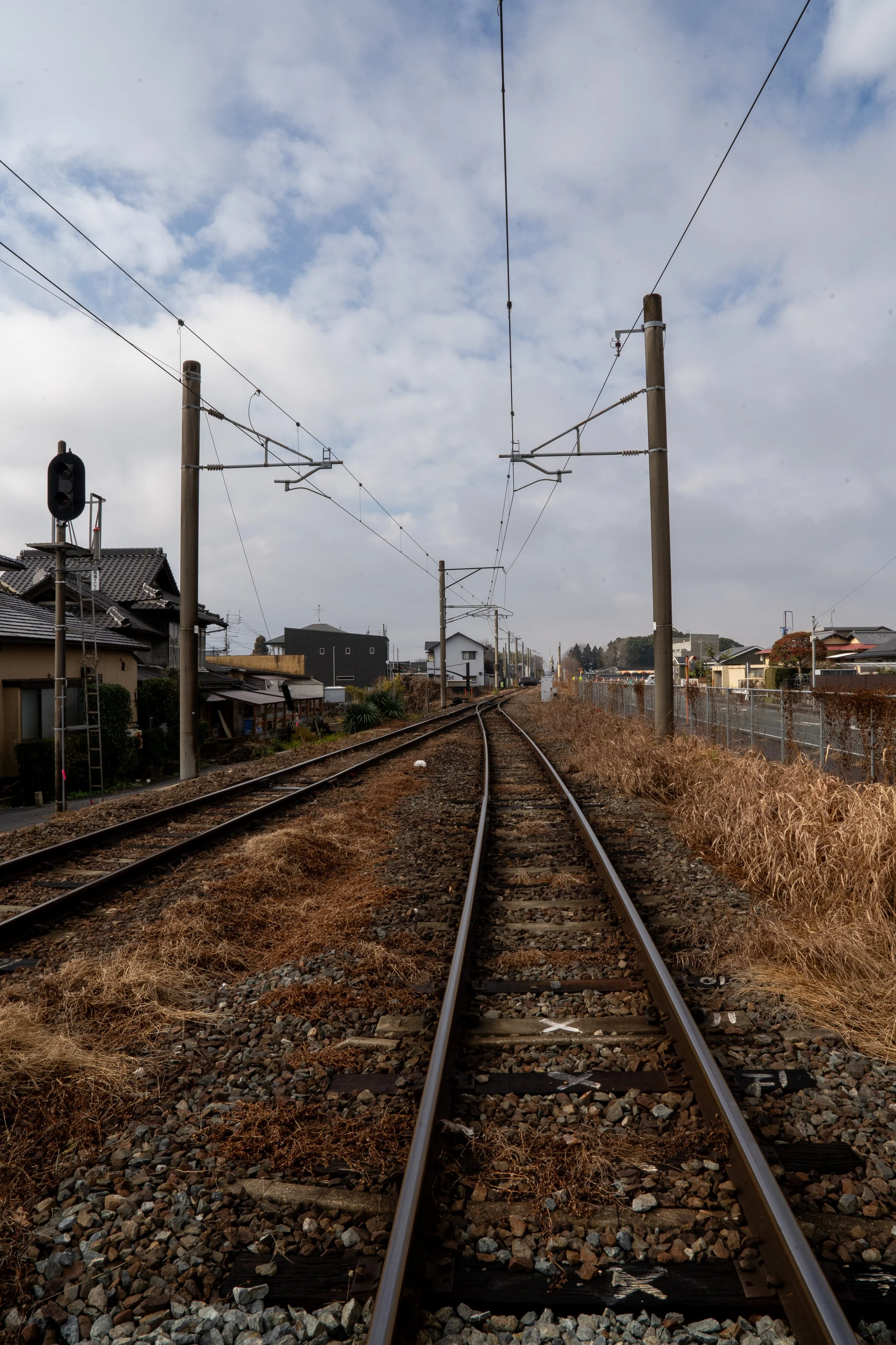 View of railway tracks stretching into the distance, with old houses and utility poles on either side, under a partly cloudy sky.