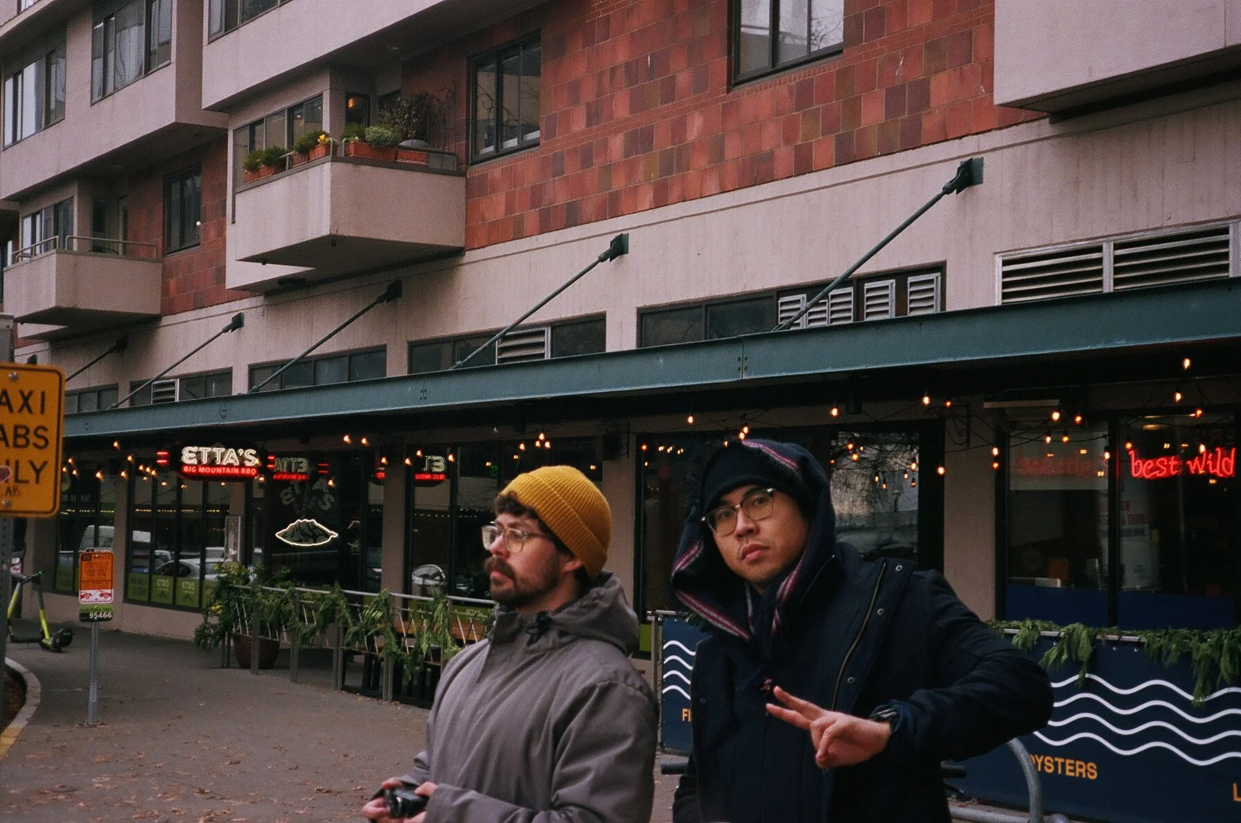 Two men standing outside a restaurant or bar in an urban area during the daytime. The man on the left is wearing glasses, a brown beanie, and a gray windbreaker, looking to the side. The man on the right is wearing glasses, a black jacket, a hoodie, 