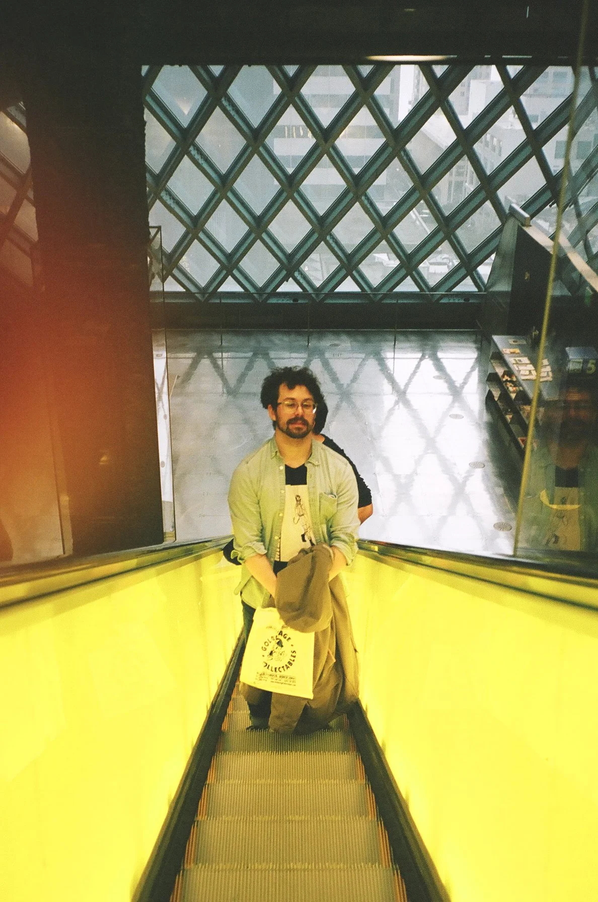 Man with glasses and beard riding an escalator with yellow sides in a modern building, holding a coat and a shopping bag, with a patterned glass window above.