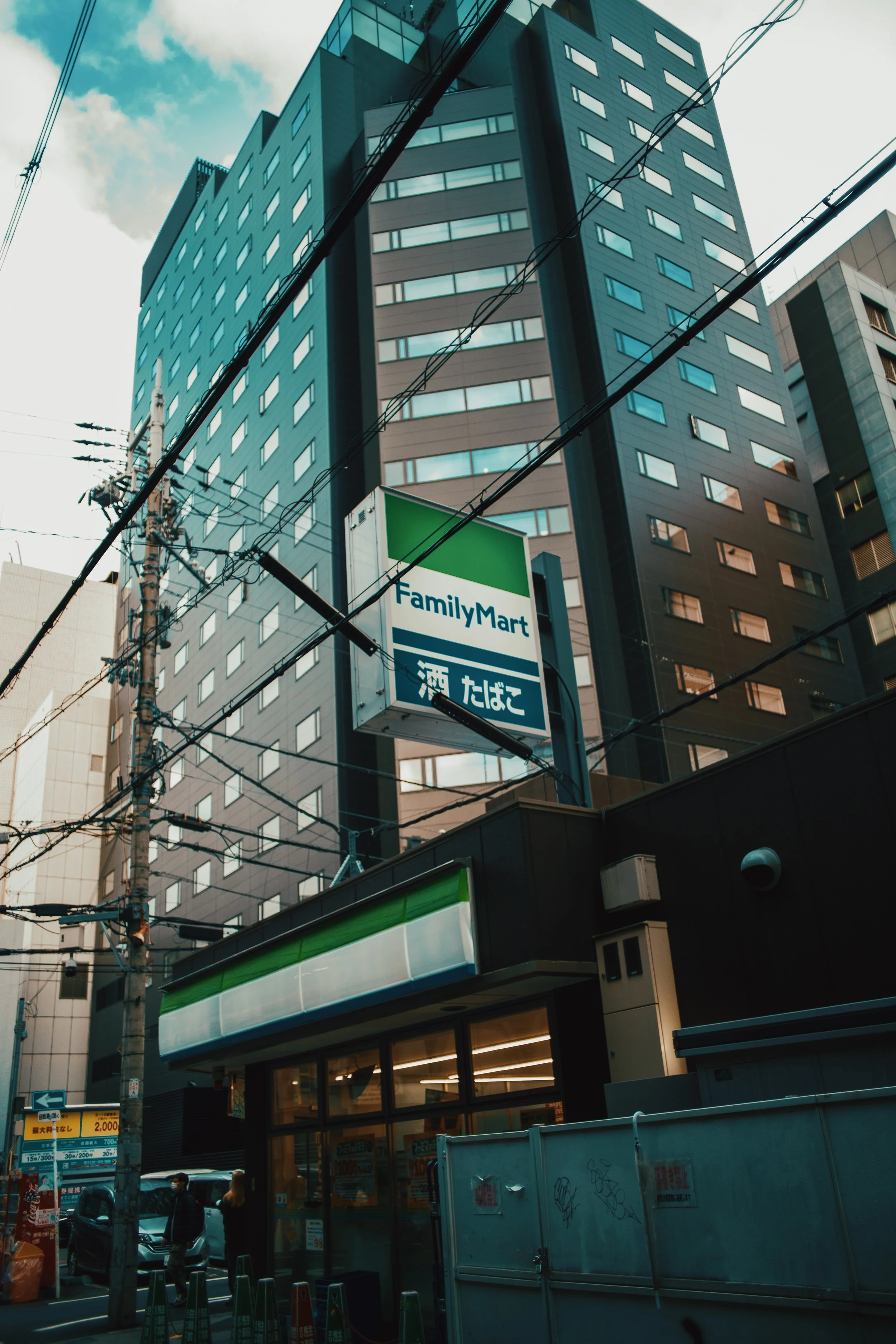 Tall modern building with a FamilyMart convenience store sign in front, city street scene with utility poles and people walking, cloudy sky above.