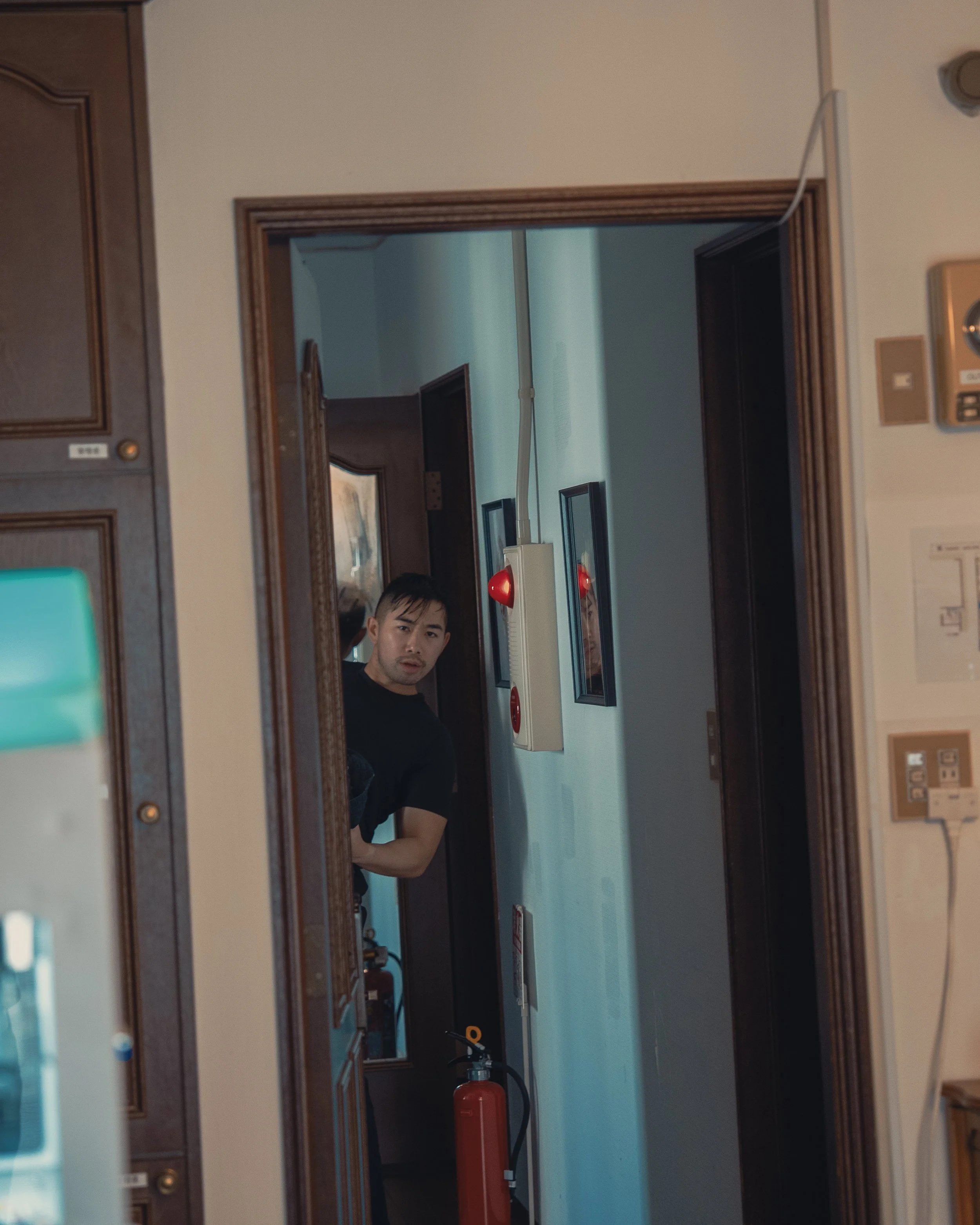 A man peeking into a room through a mirror in a kitchen with wooden cabinets, wall-mounted fire alarm, fire extinguisher, framed pictures on the wall, and various electrical outlets.