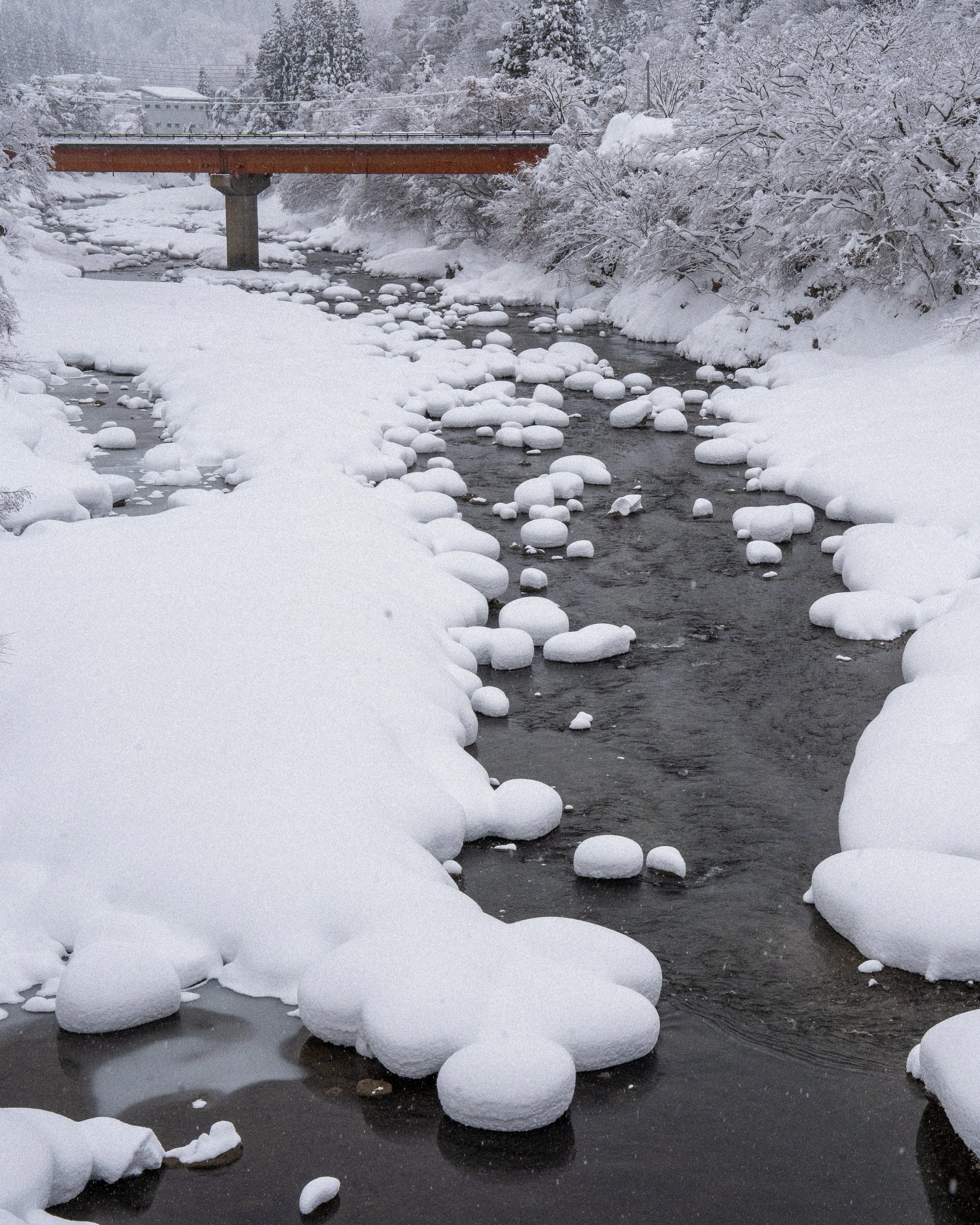 A river flowing through a snow-covered landscape with a bridge in the background, surrounded by snow-laden trees and rocks.