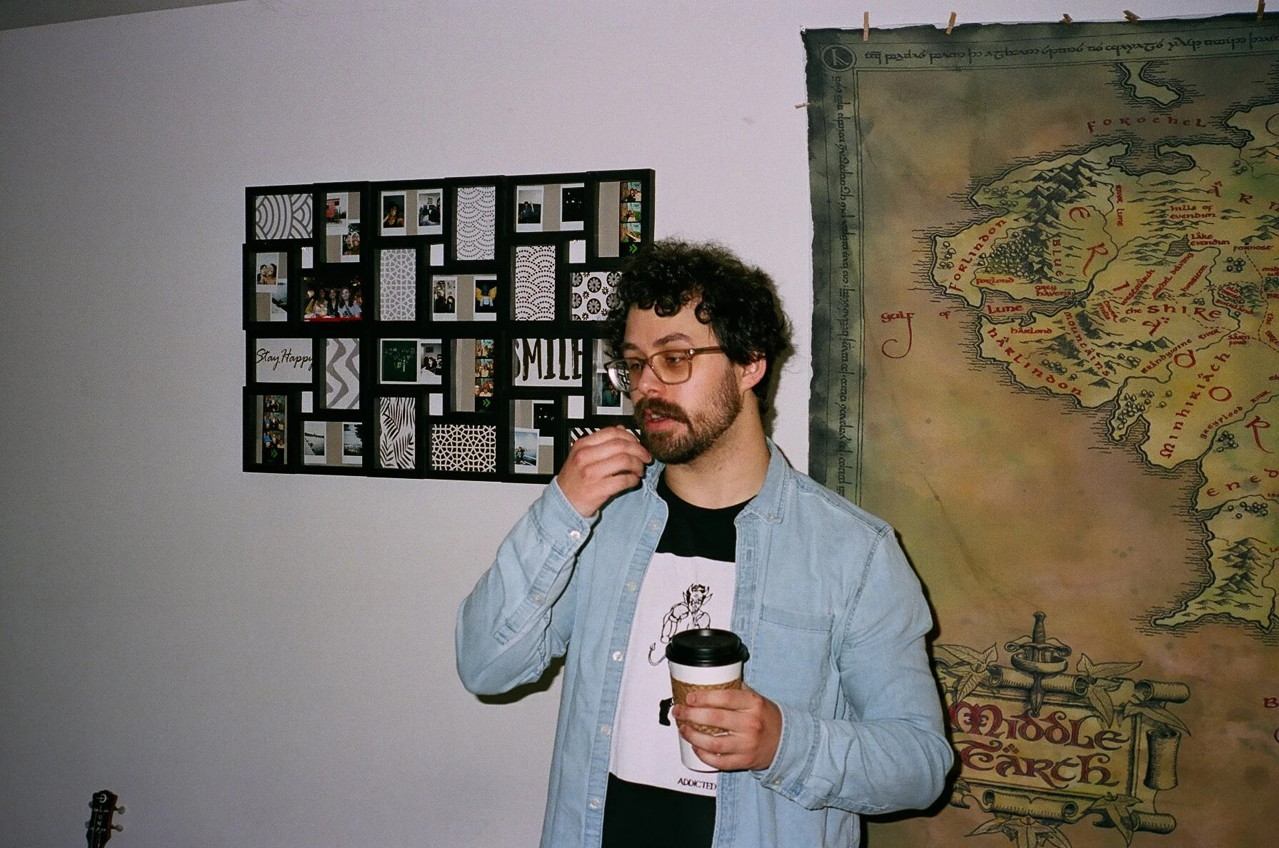 A man with glasses and a beard holding a coffee cup, wearing a light denim jacket and a graphic T-shirt, standing in front of a collage of photos on the wall and a large illustrated map.