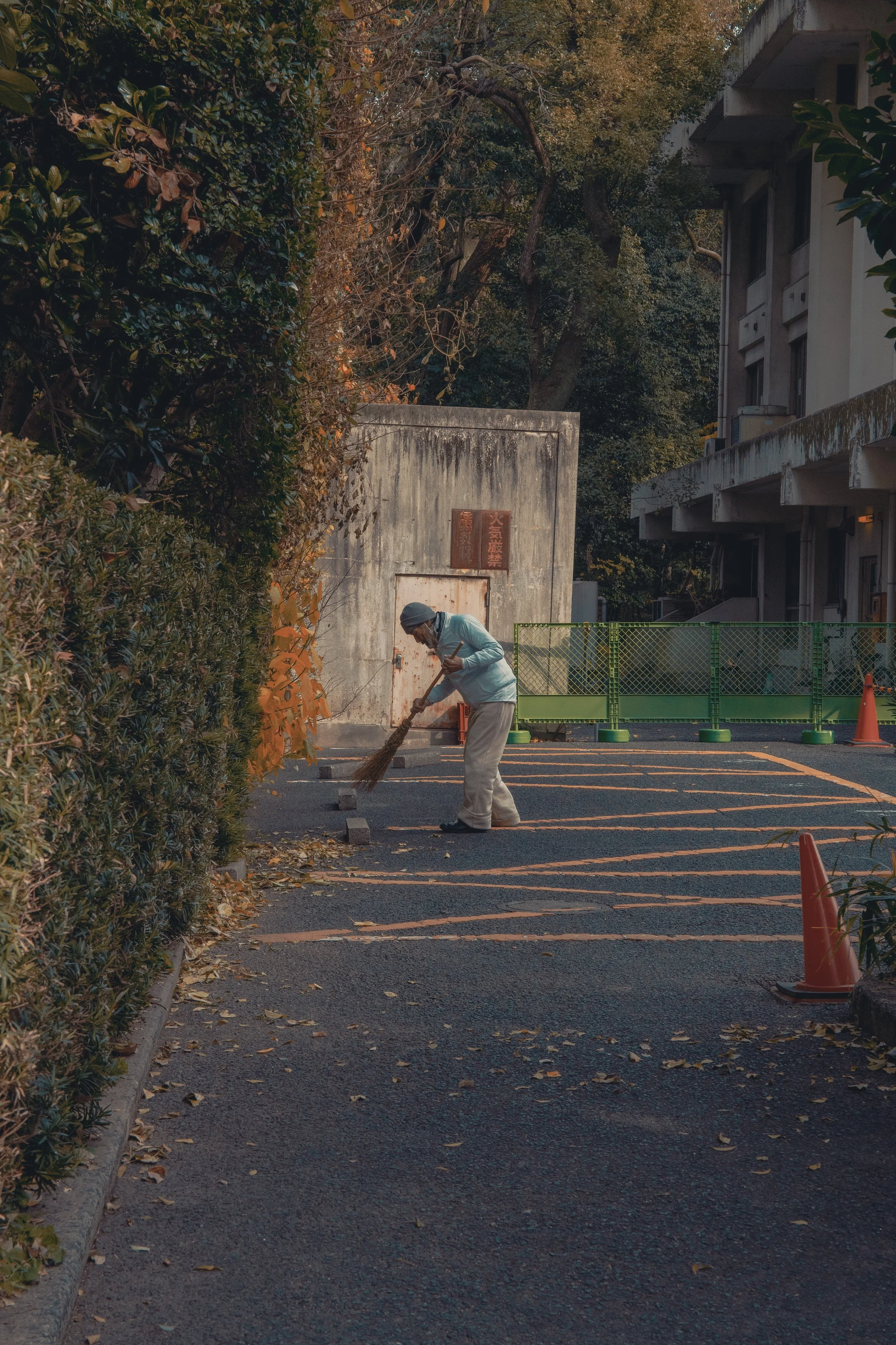 A person sweeping fallen leaves in an outdoor parking lot with barriers and trees in the background.