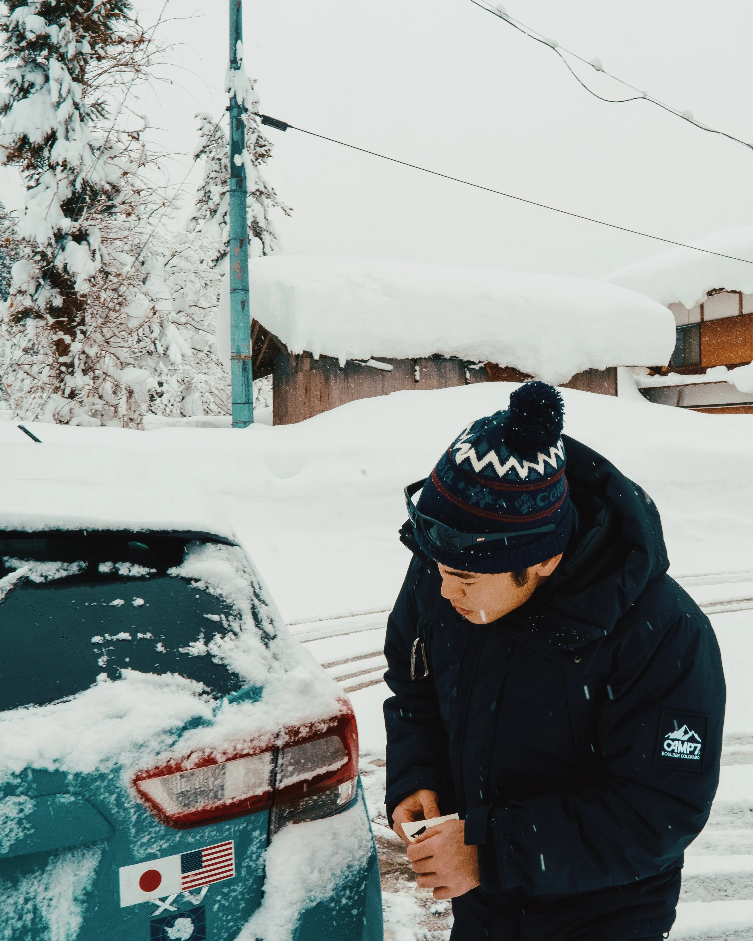 A person dressed in winter gear, including a black jacket with a Camp 7 patch and a blue knit hat with a pom-pom, stands next to a snow-covered car in a snowy landscape. There are snow-covered trees, a wooden structure, and utility wires in the backg