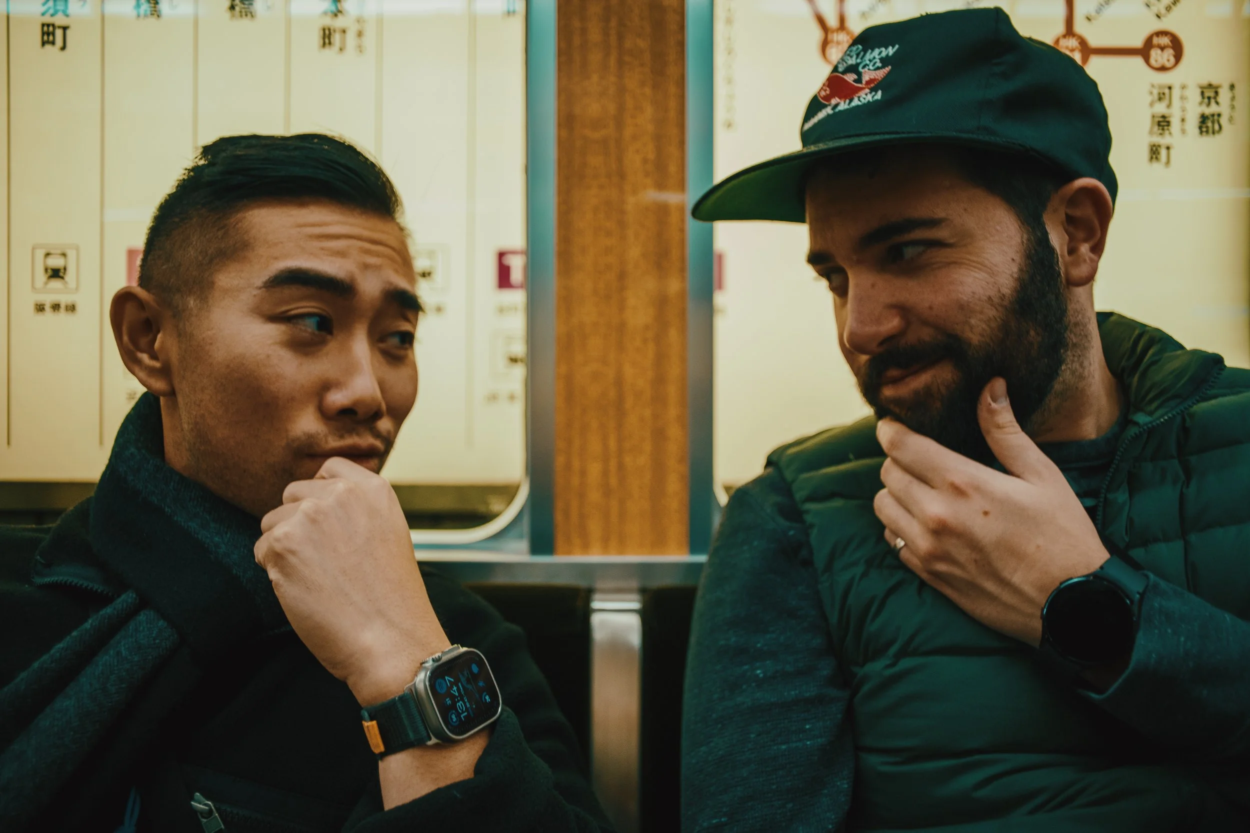 Two men in a train station sitting and having a conversation, with Japanese signs on the wall behind them.