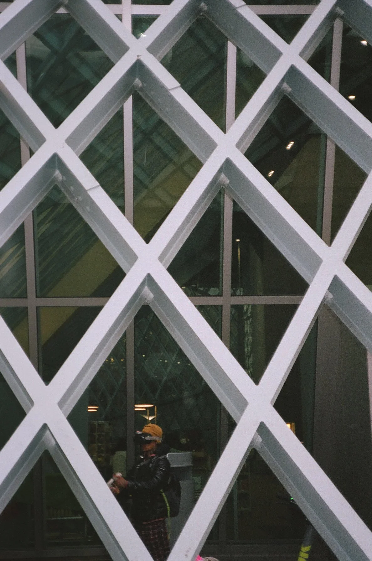 Person standing behind a decorative lattice wall outside a glass building, looking at their phone, wearing a yellow hat and a leather jacket.