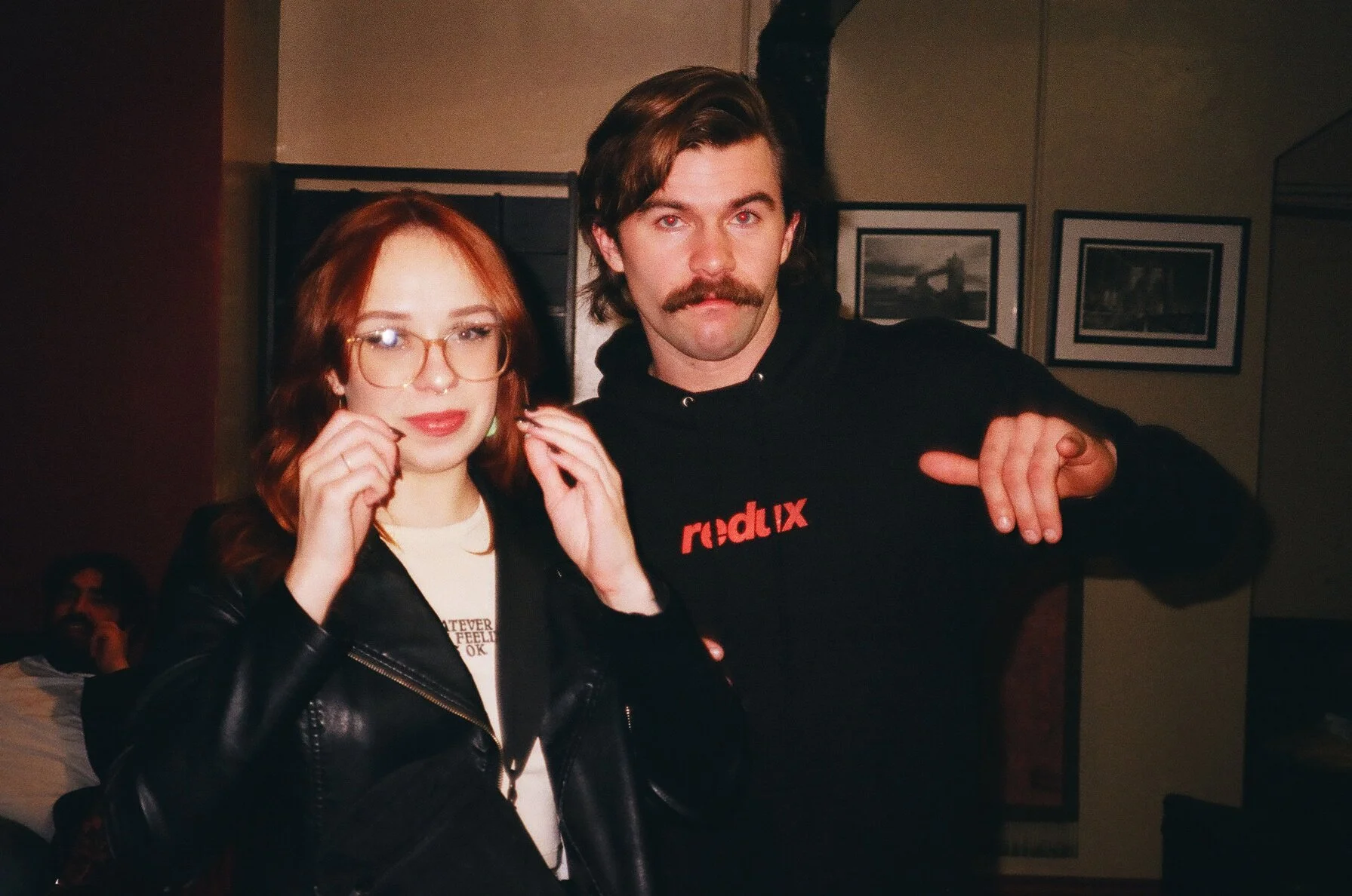 A young woman with red hair, glasses, and a black leather jacket standing next to a man with a mustache and a black hoodie that has red text. They are posing for the camera indoors, with framed pictures hanging on the wall in the background.