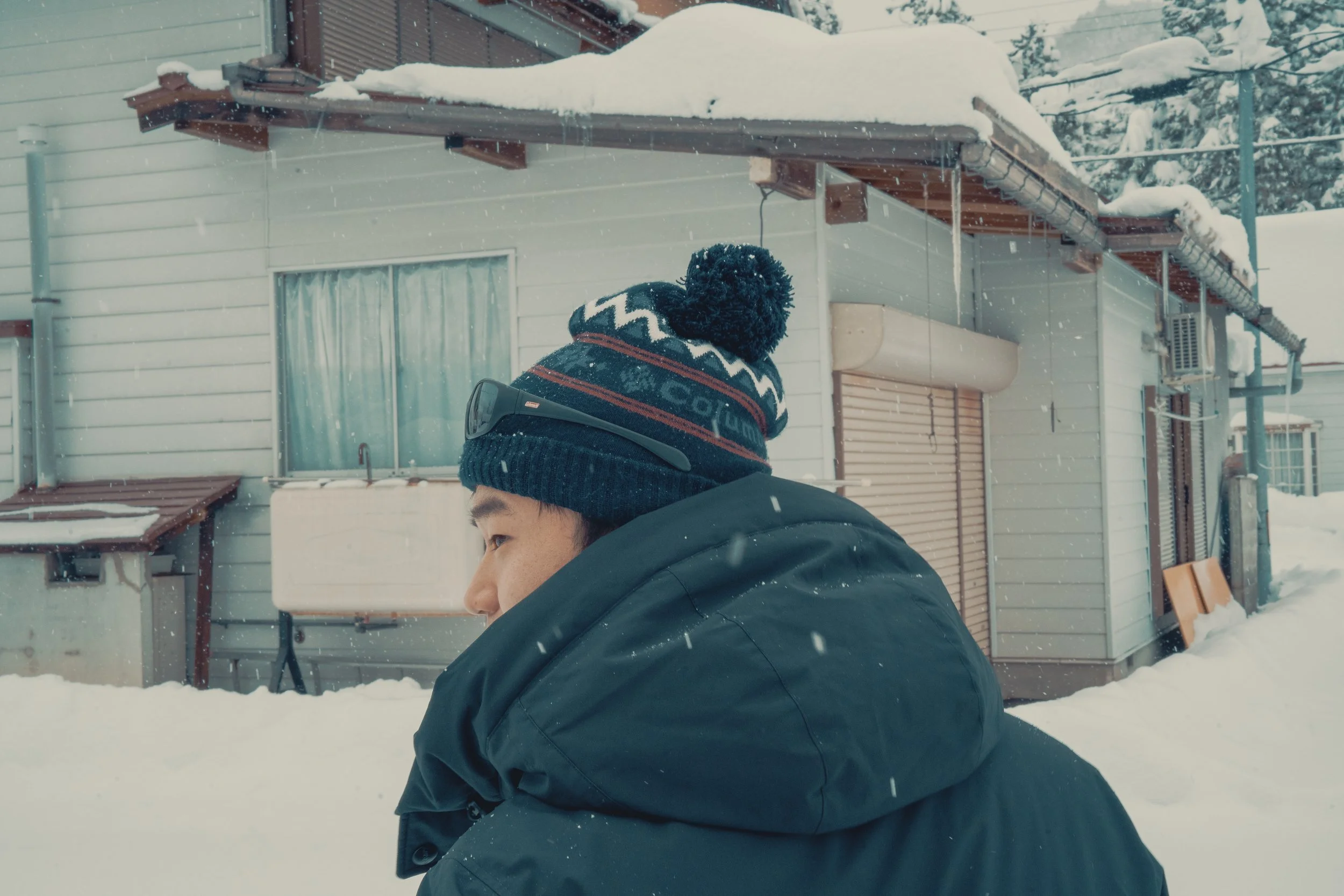 A person wearing a winter coat and a blue and red pom-pom beanie standing outside in snow, with a house in the background.