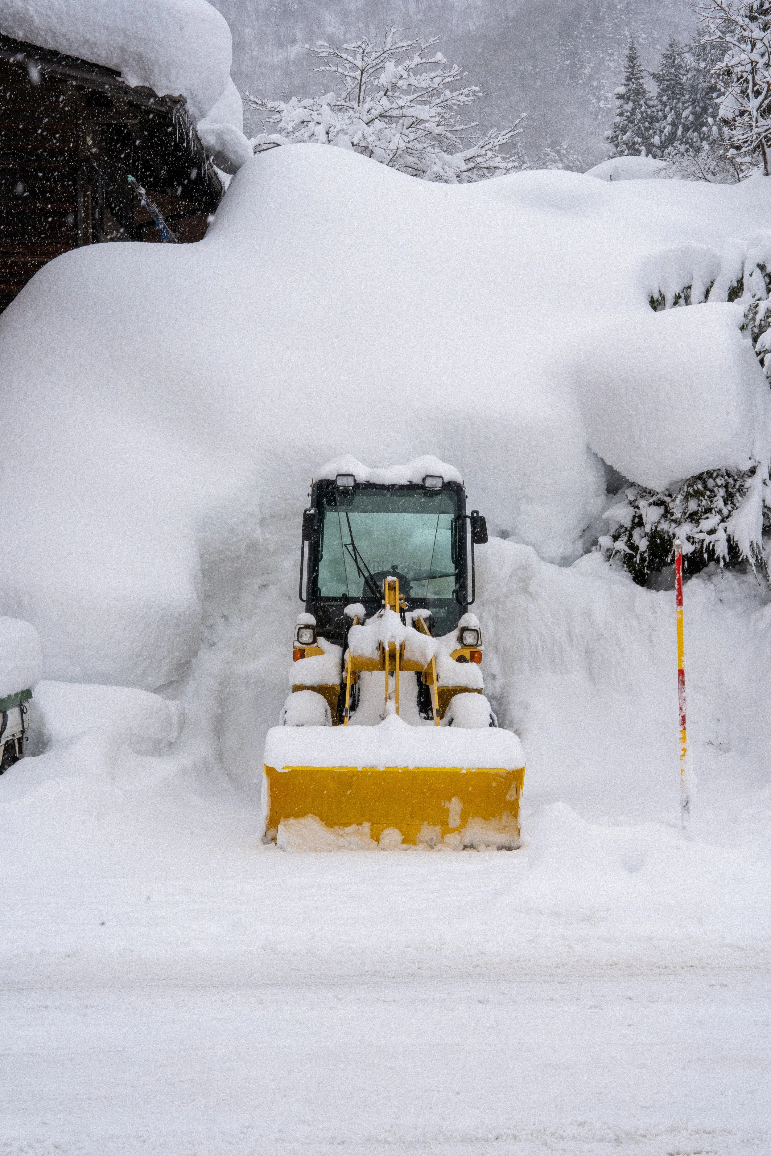 A snow plow clearing a pathway through deep snow in a winter landscape with snow-covered trees and a building in the background.