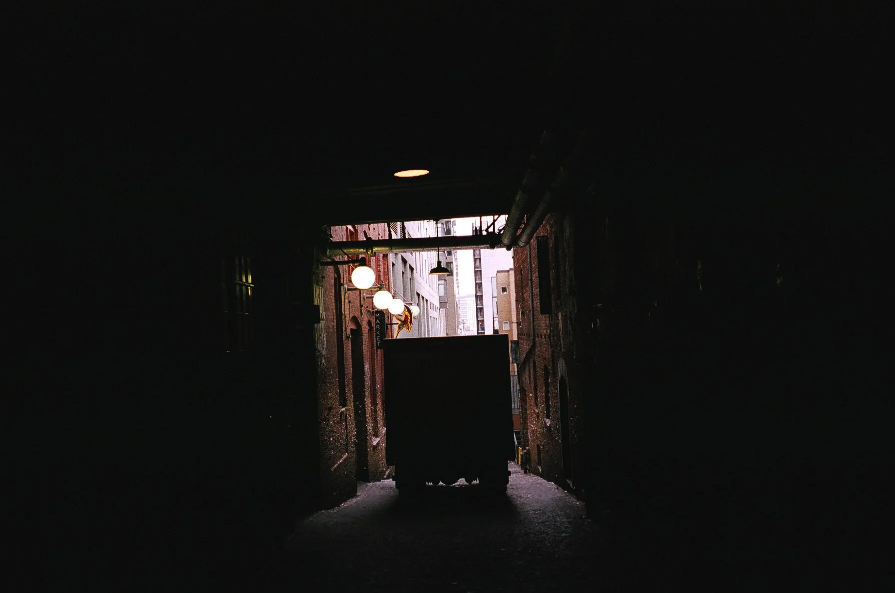 Dark alleyway with a large trash bin and illuminated street lights in the background.