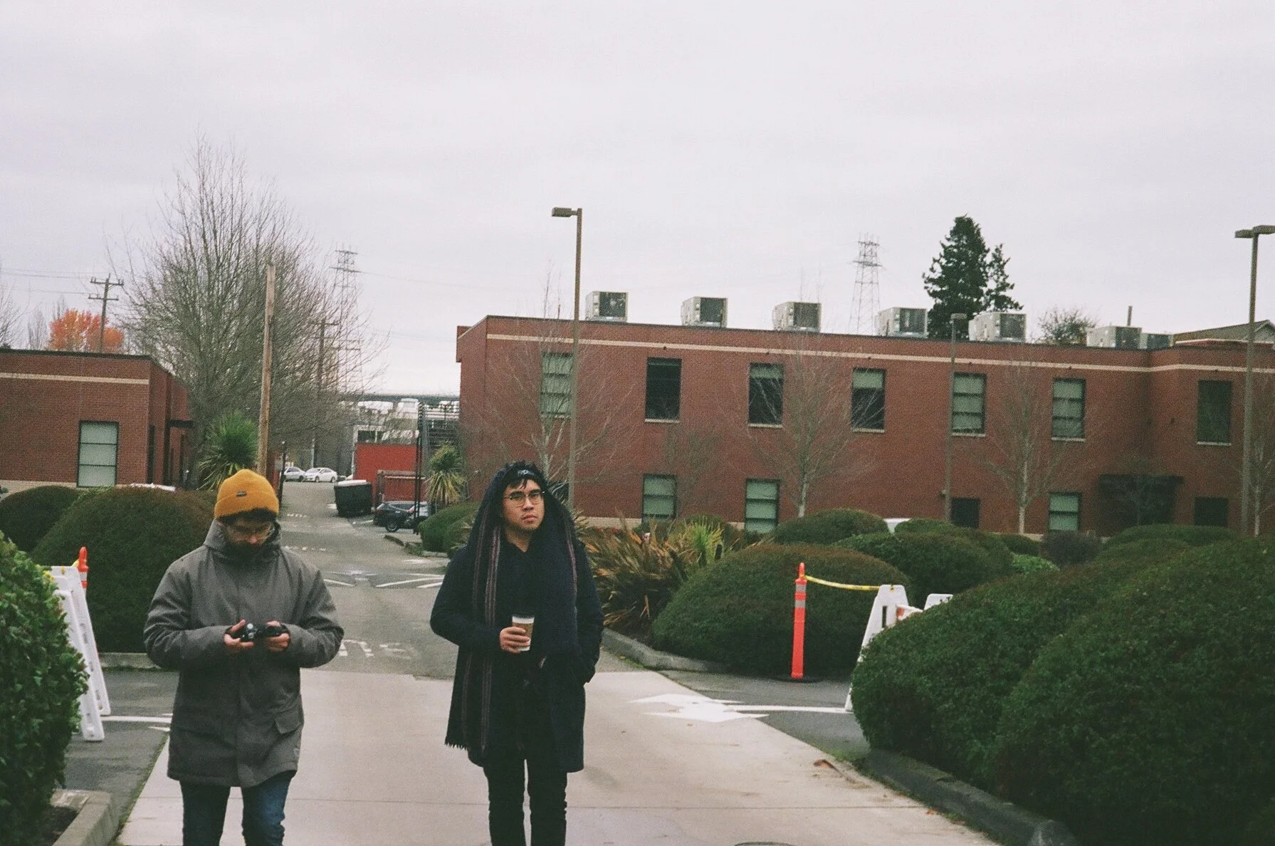 Two people walking along a sidewalk in a parking lot with bushes and brick buildings in the background on an overcast day.