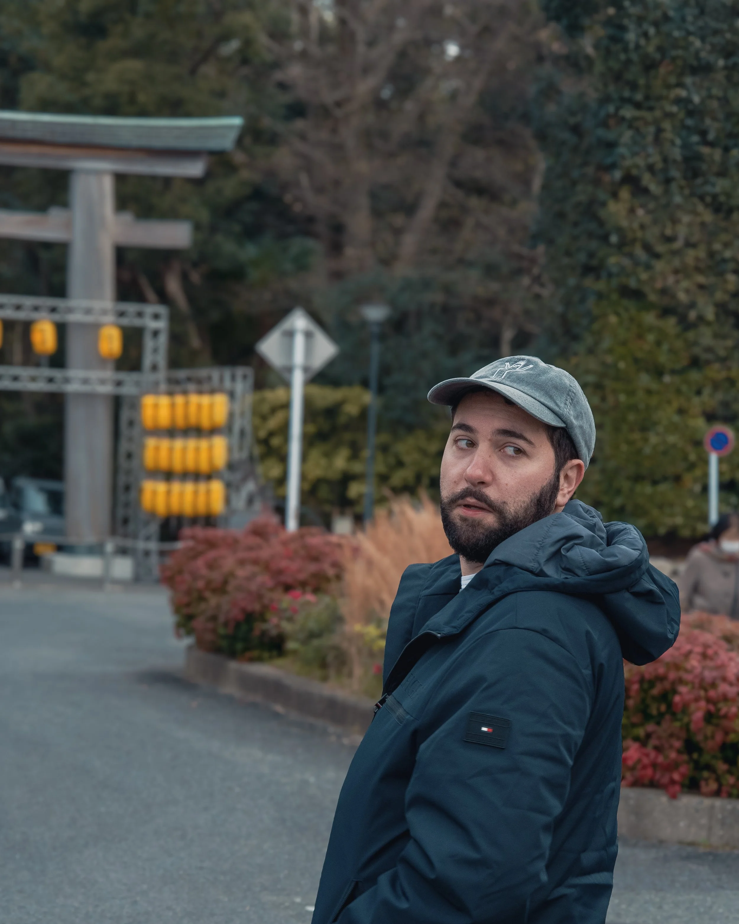 A man with a beard, dark hair, and a gray cap looking over his shoulder outdoors near a cultural site with a torii gate and autumn foliage.