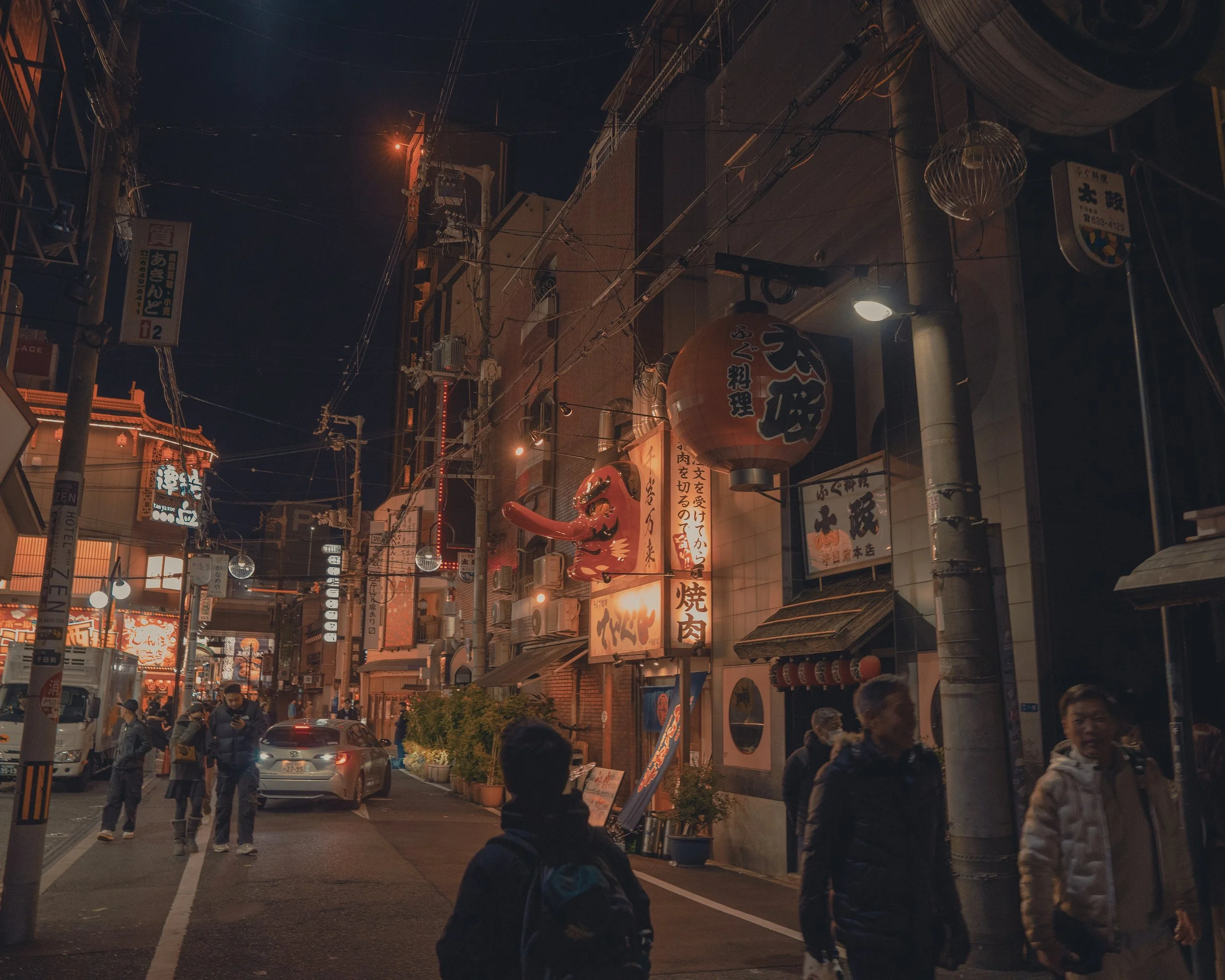 Nighttime scene of a busy Japanese street with illuminated signs, lanterns, and pedestrians.