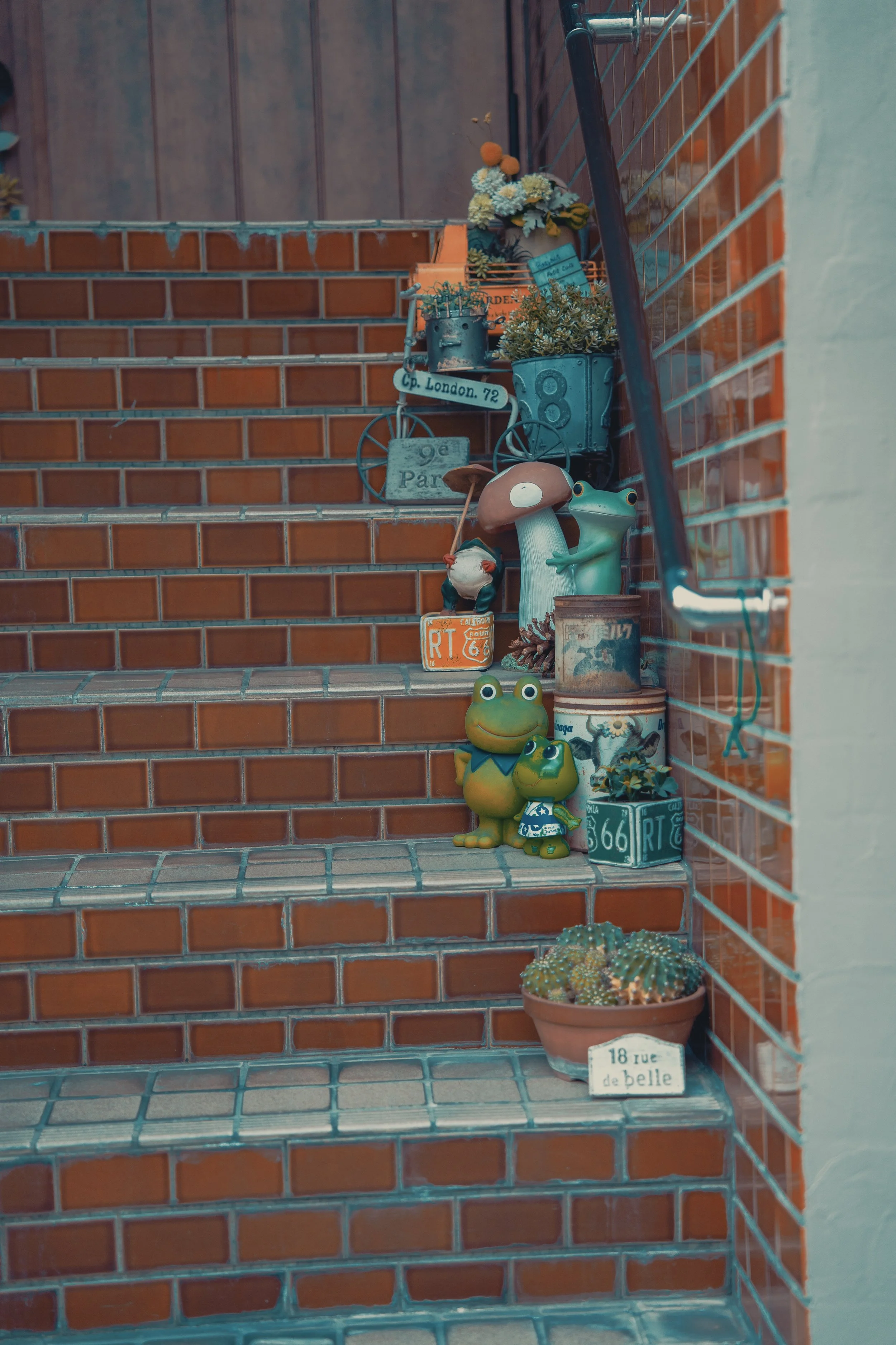 Decorative plants, frog figurines, and vintage cans arranged on brick stairs near a brick wall.