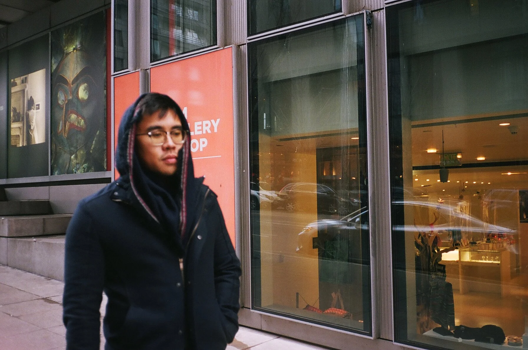 A person with glasses and wearing a dark jacket walking past a gallery shop with large glass windows on a city street.