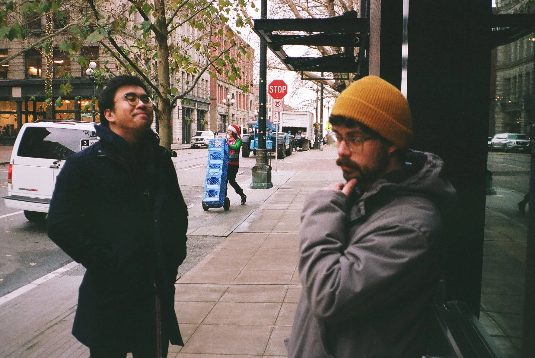 Two men standing on a city sidewalk, conversing, with trees, parked cars, and a woman in a Santa hat in the background.
