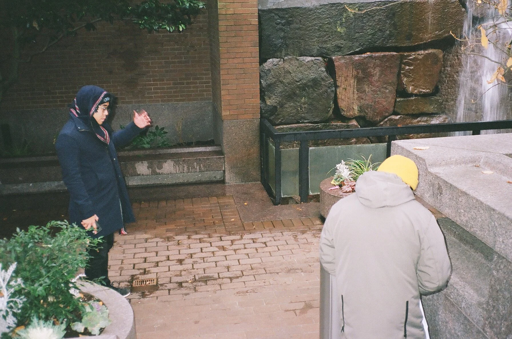 A person in a beige jacket and yellow hat bowing in front of a person dressed in dark clothing and a hoodie in an outdoor urban setting with brick pavement, plants, and stone walls.