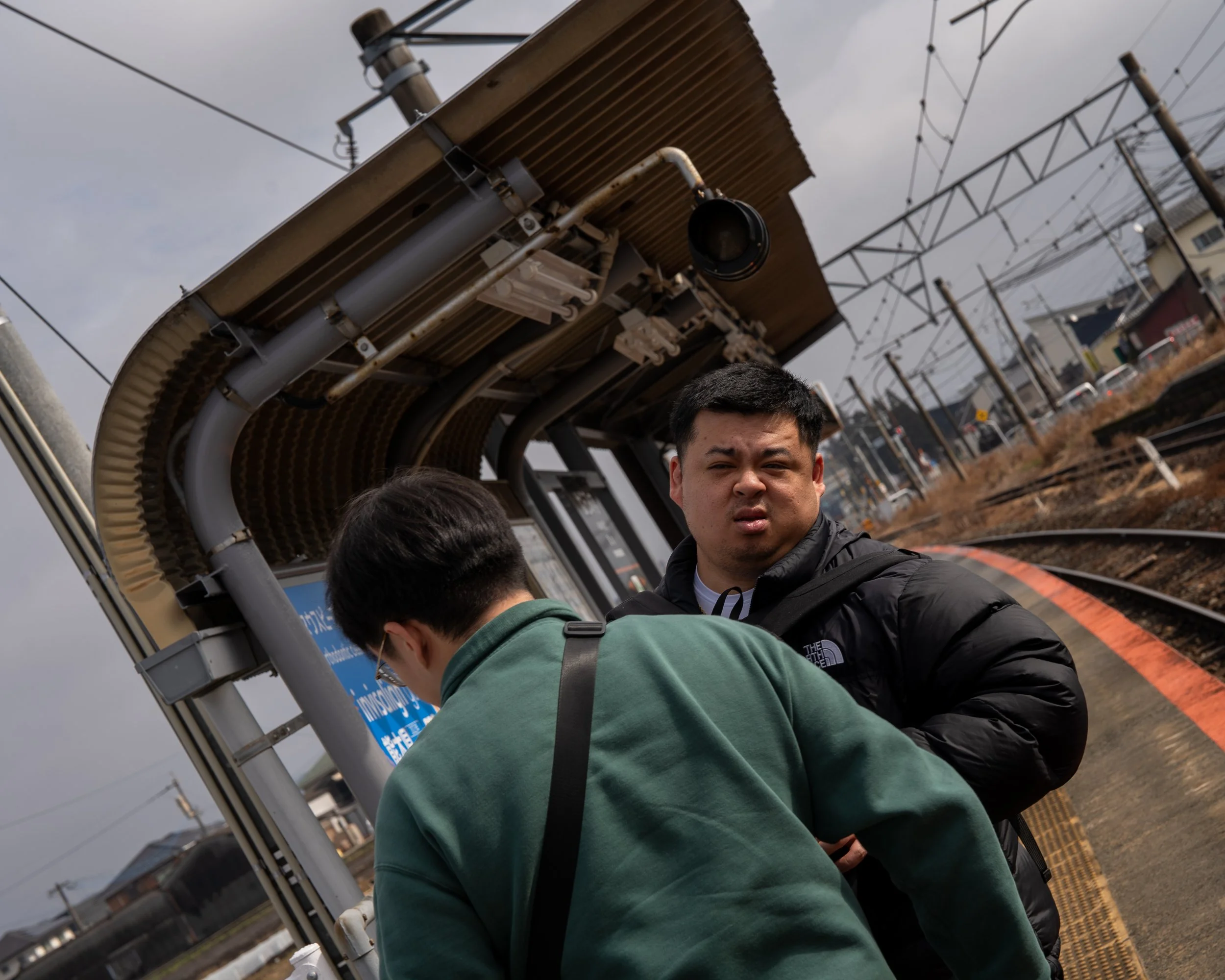 Two young men stand on a train platform beneath a shelter, one facing away with a green jacket and the other facing the camera with a black jacket, looking confused, with train tracks and power lines in the background.