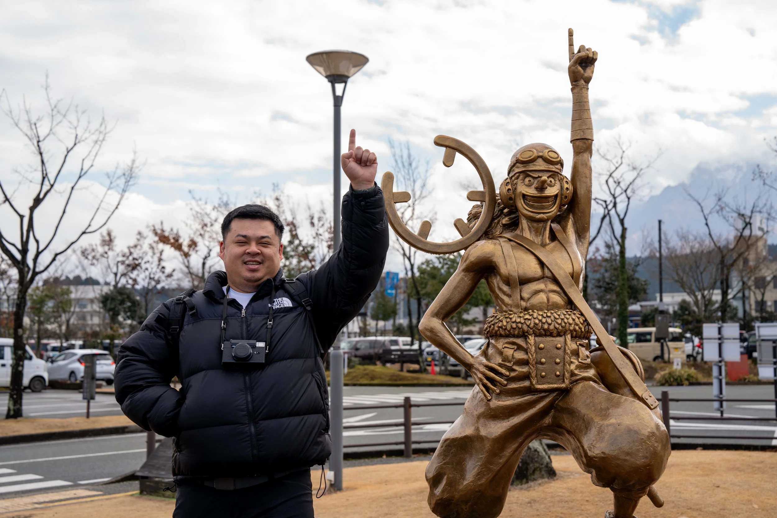 A man in a black jacket with a camera around his neck stands next to a large bronze statue of a character with a big smile, goggles, and a wide stance, with one arm raised pointing upward in a gesture of enthusiasm. The scene is outdoors with trees, 