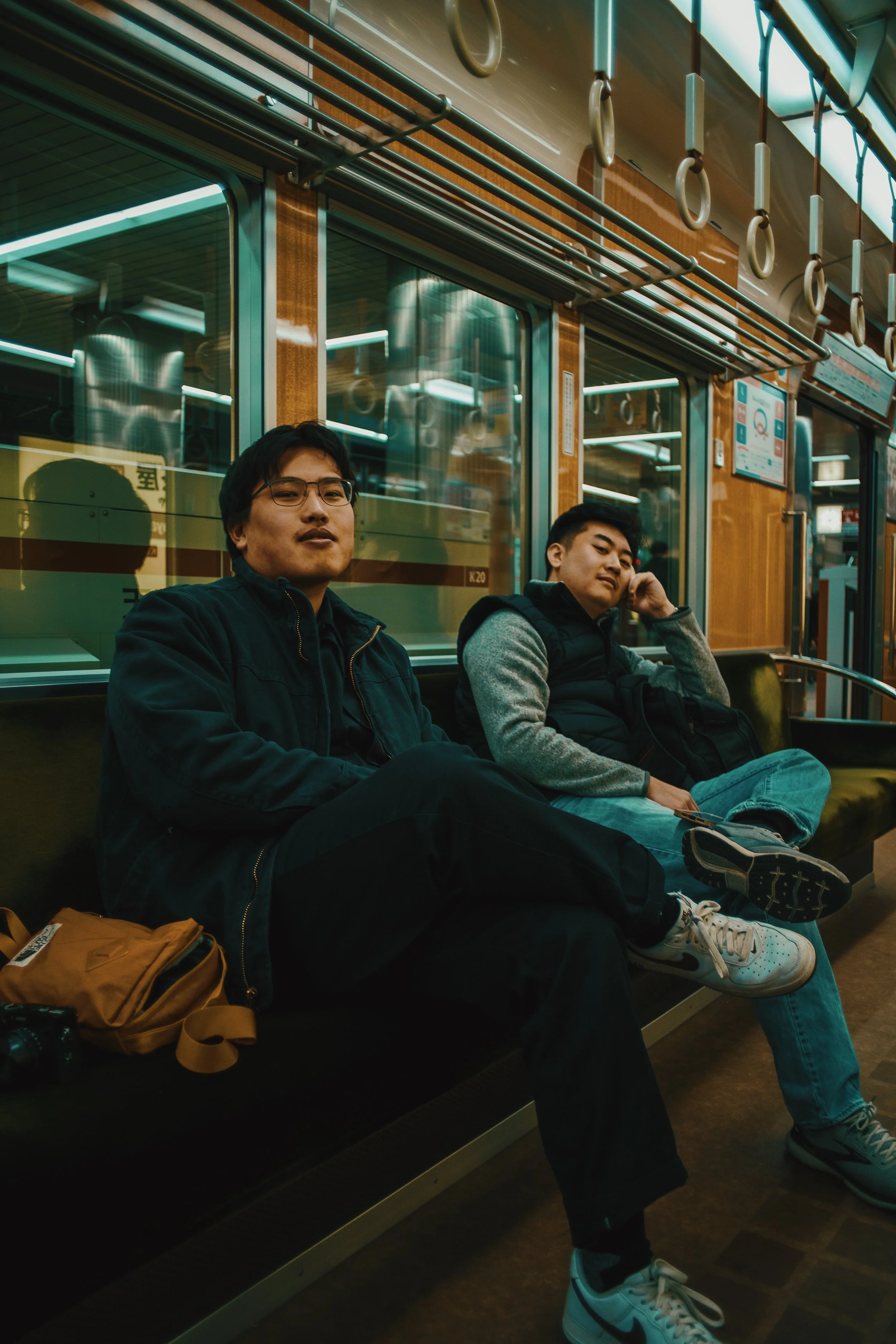 Two young men sitting on a subway train, one wearing glasses with black hair, the other resting his head on his hand, both dressed in casual clothing, with bags beside them, inside a subway car with reflective windows and handrails.