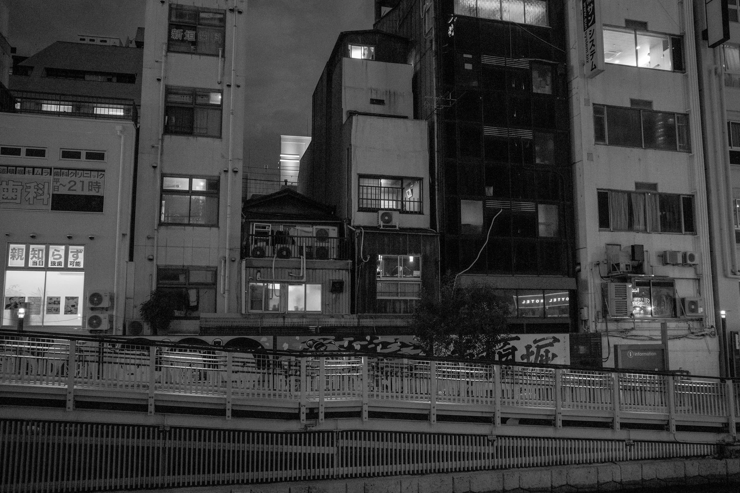 Nighttime cityscape of tall, closely packed buildings with illuminated windows and signs, viewed from across a river with a railing in the foreground.
