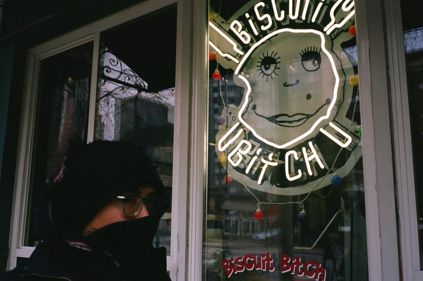 Person wearing glasses and a black hooded jacket standing outside a store with a neon sign that reads 'Biscuit Bitch' and features a cartoon girl face with freckles and eyelashes.