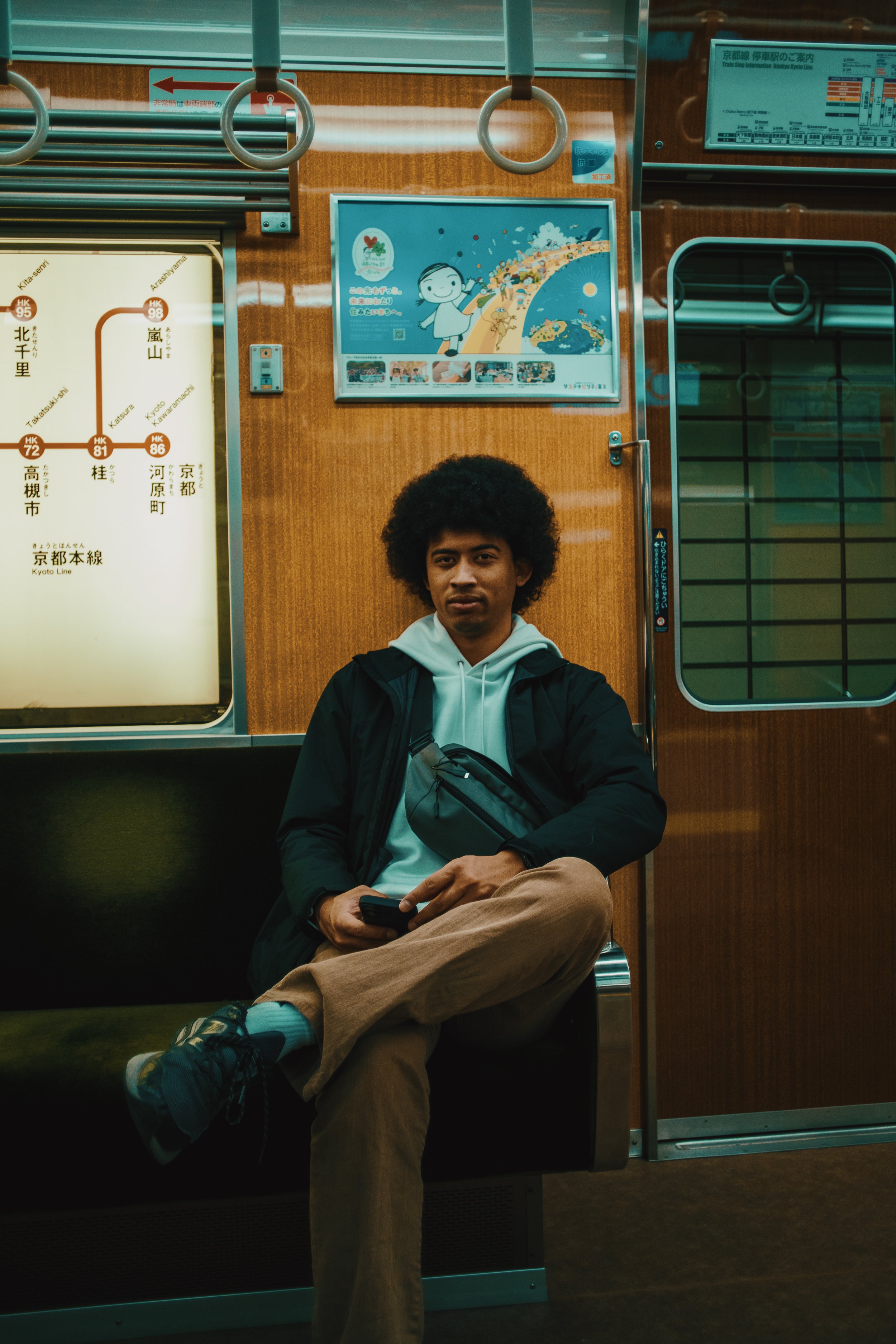 A young man with curly hair sitting inside a train, holding a phone, wearing a white hoodie, black jacket, beige pants, and sneakers. The train has wooden paneling and a map with Japanese characters and signs around him.