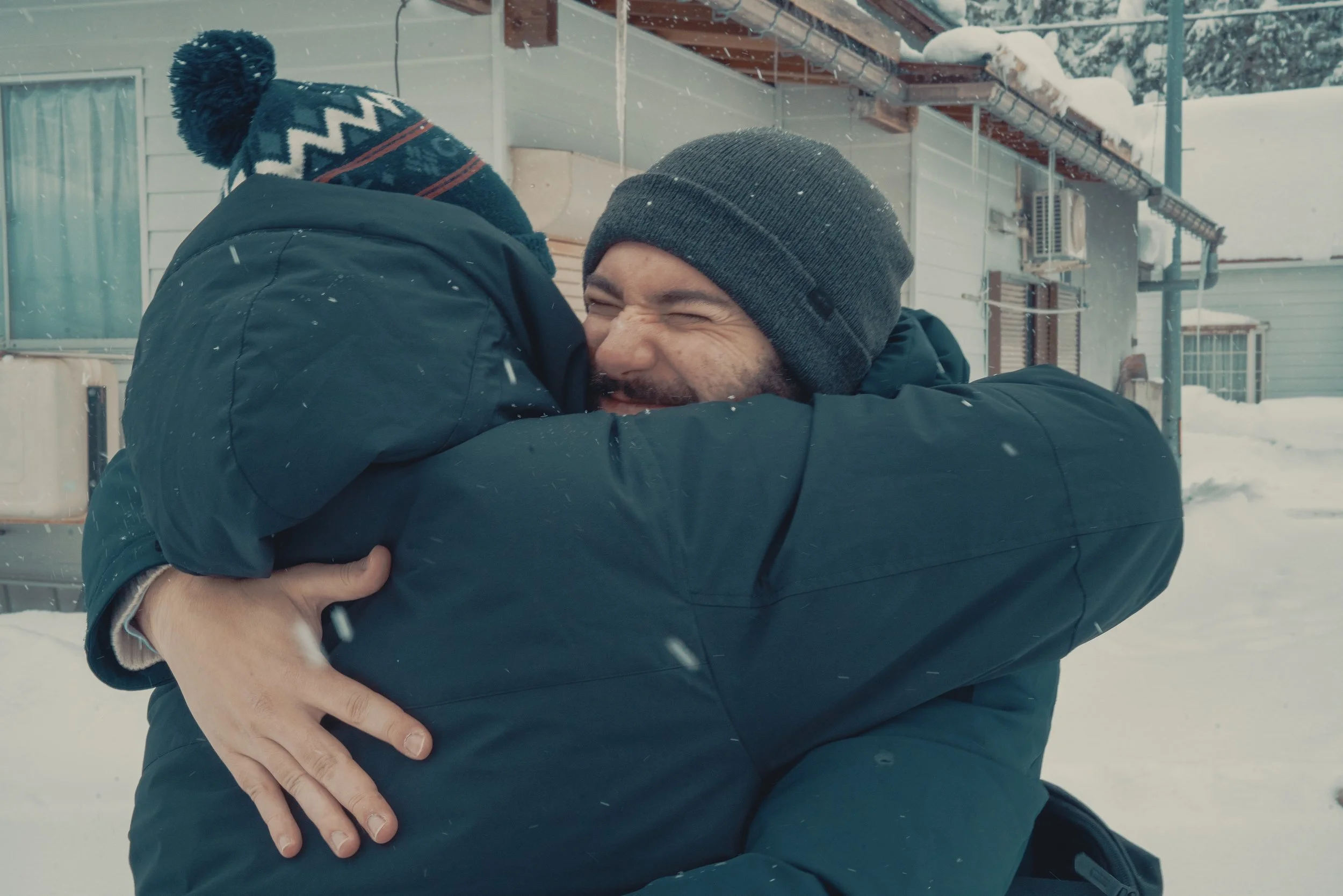 Two men wearing winter clothes hugging warmly outside in a snowy environment, showing joyful emotion.