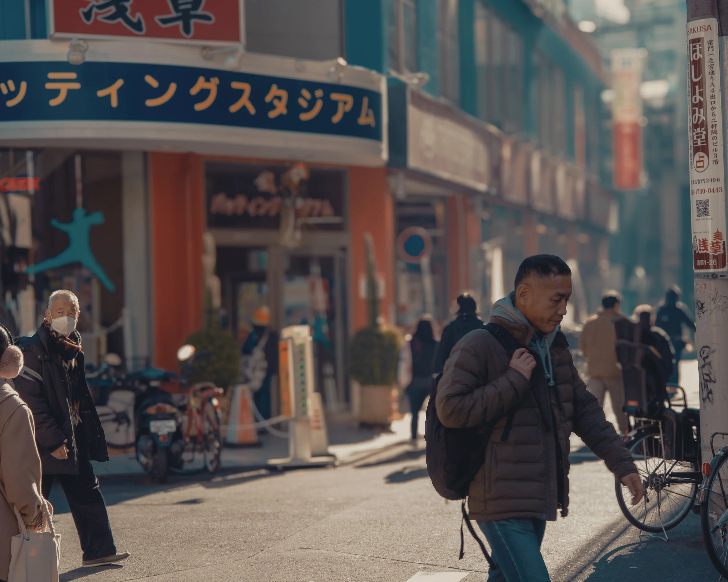 People walking on a busy street with storefronts, bicycles, and Japanese signs, with some wearing masks.