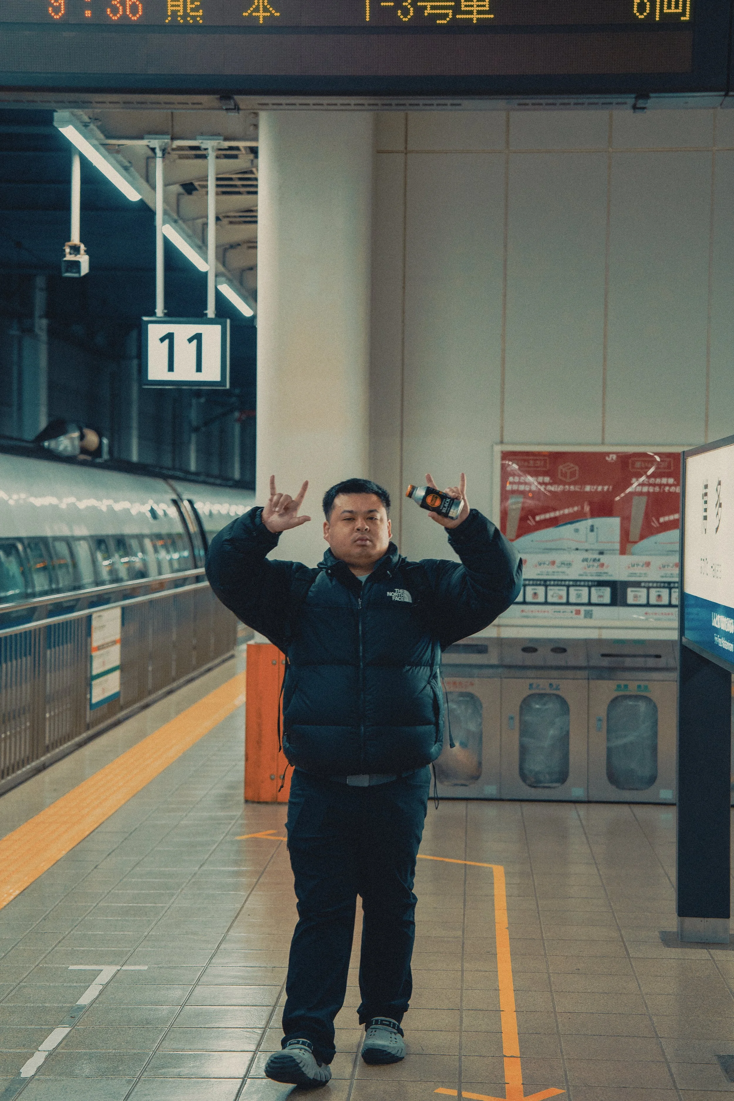 A man standing on a subway platform, making rock and roll hand signs with both hands, holding a beverage in his right hand. The platform is empty, and there are vending machines in the background. An overhead sign displays the number 11.