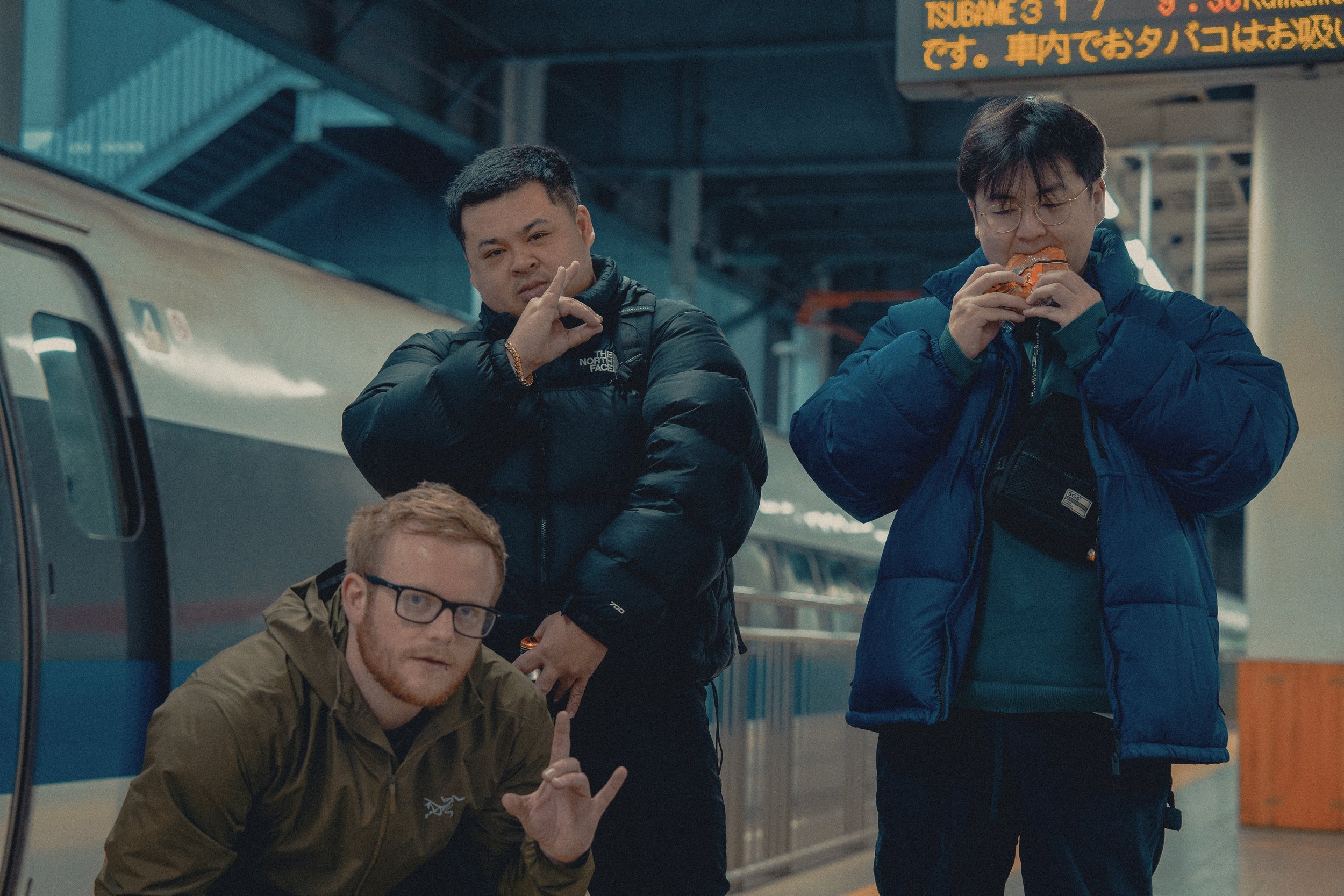Three men in winter jackets at a train station. One man is crouching and making a gesture with his hand, another is standing with glasses eating a sandwich, and the third man is standing with his hand on his chin, posing. A train is visible in the ba