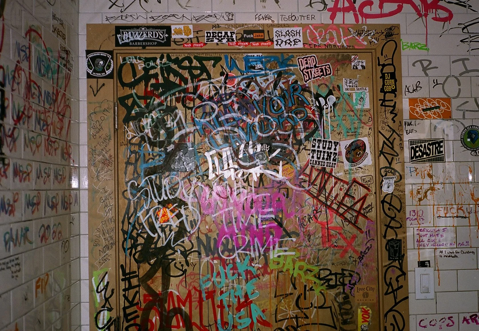 A wall and door covered with various colorful graffiti, stickers, and writings in different styles and sizes.