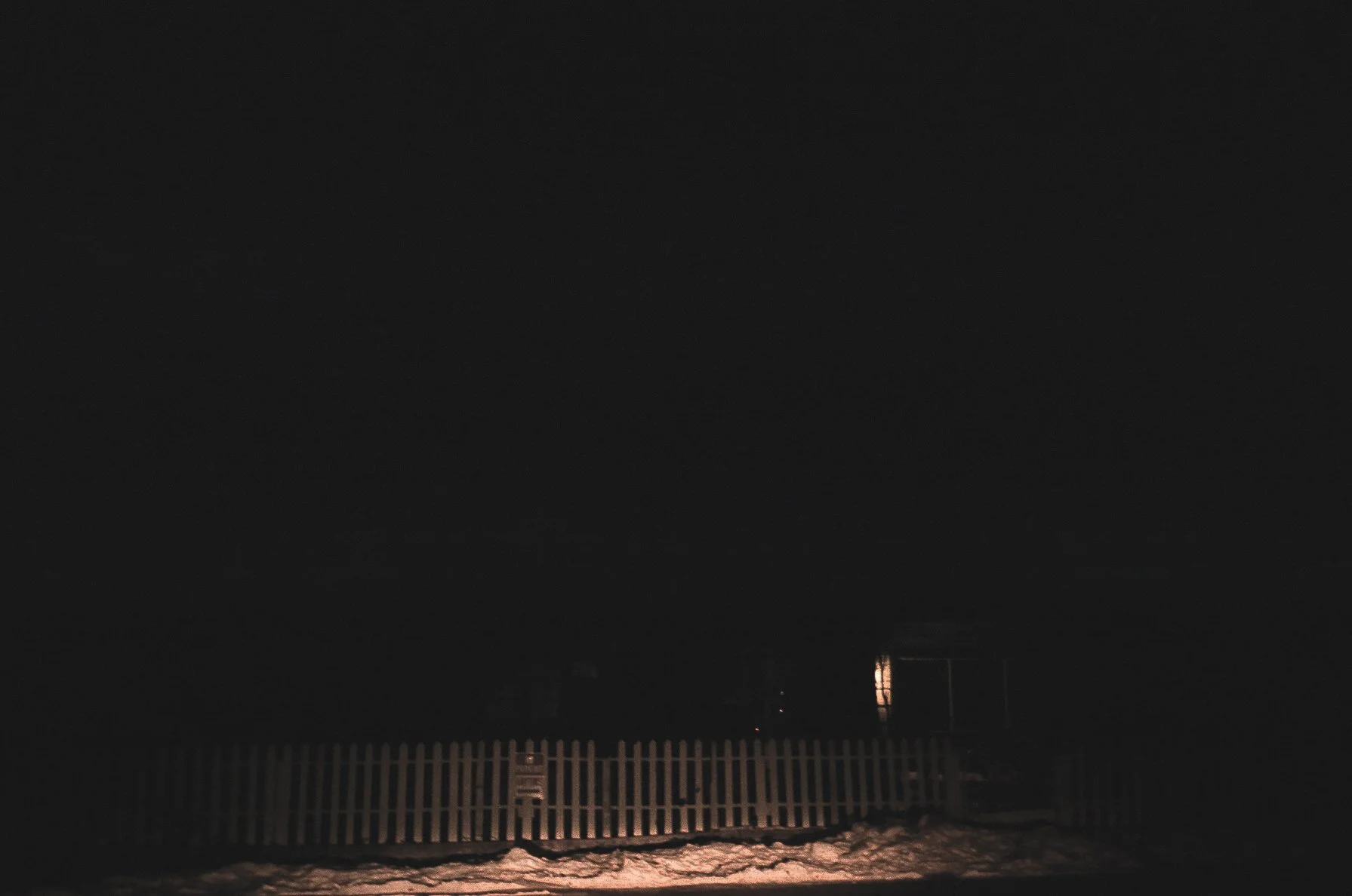 Dark night scene with a snow-covered ground and a wooden fence, with a faint glow from a distant light in the background.