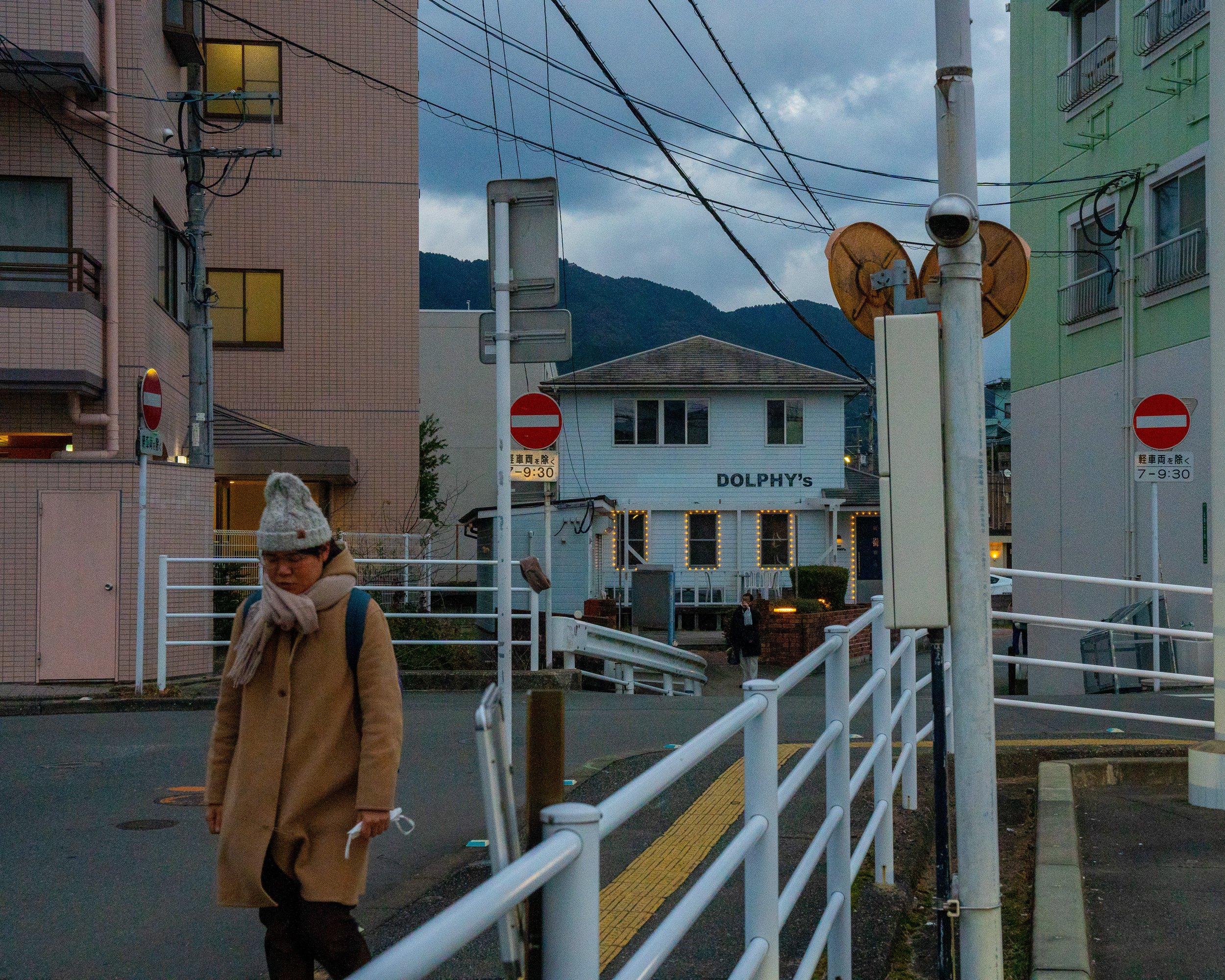 A woman in a beige coat, scarf, and gray knit hat walks past a white railing with yellow tactile paving on the sidewalk, with residential buildings and a small white structure with a sign that reads 'DOLPHY's' in the background. Overhead, there are p