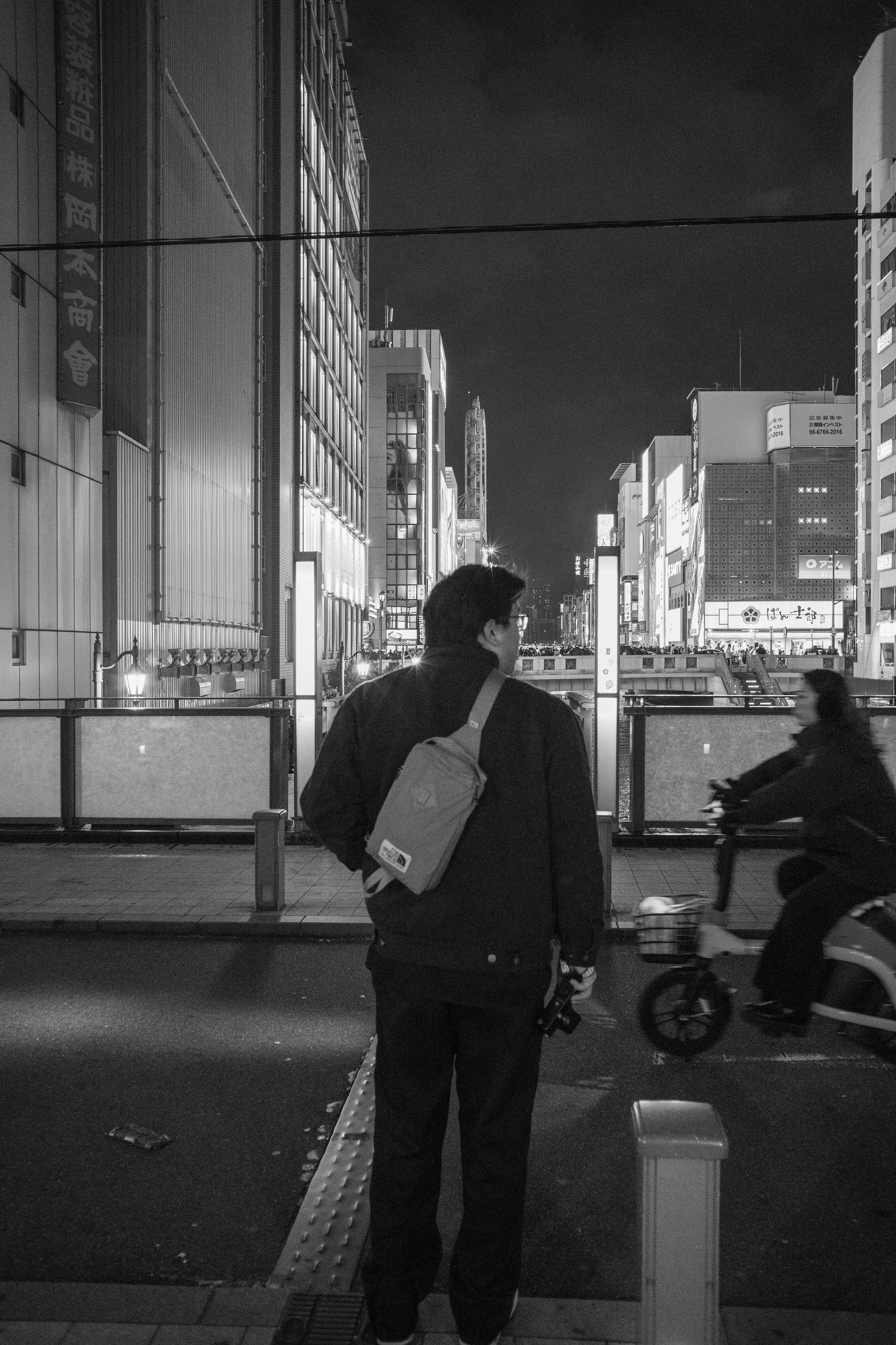 A man with a backpack stands on a city street at night, looking at a woman riding a bicycle. The background features tall illuminated buildings, suggesting an urban setting.