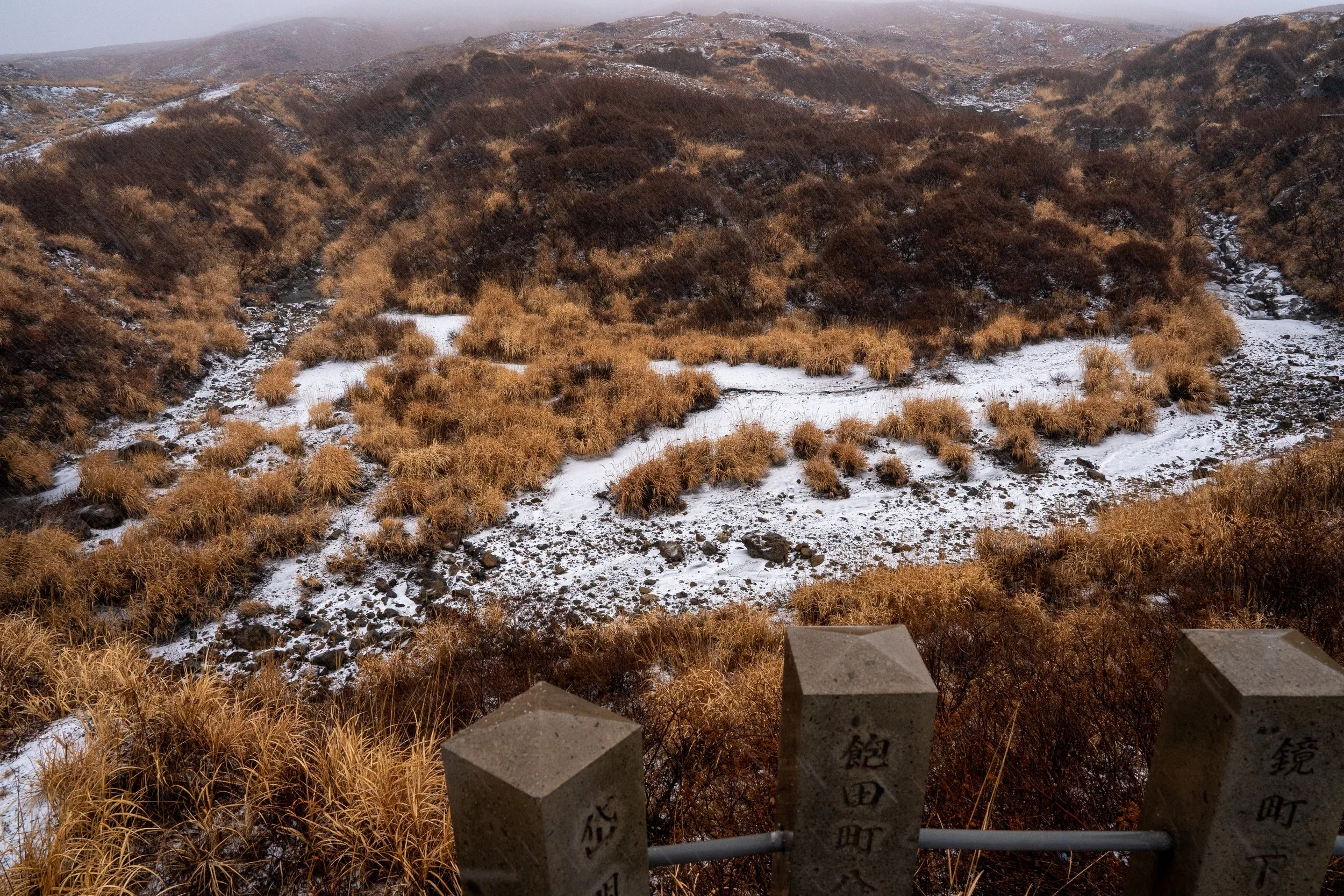 A mountainous landscape with brown and golden shrubs, patches of snow, and a cloudy sky. A concrete railing with Japanese inscriptions is in the foreground.