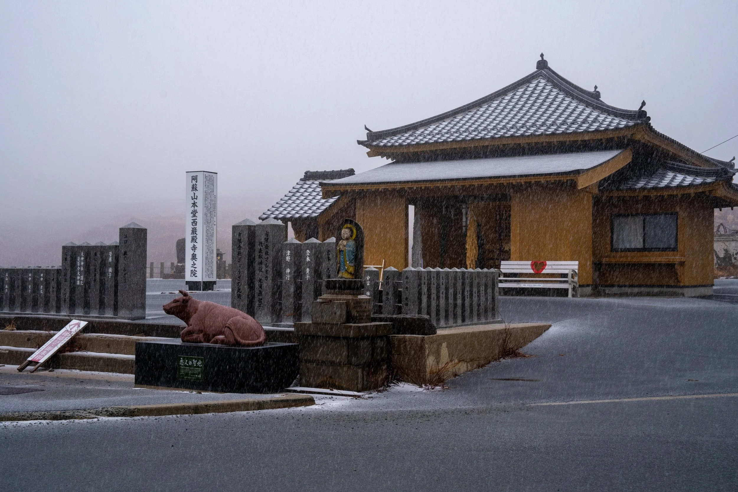 A traditional Japanese temple with a tiled roof and yellow walls, situated outdoors on a rainy day with falling rain and wet pavement, surrounded by stone statues and a small statue of a seated figure, with a white bench and Japanese signs nearby.