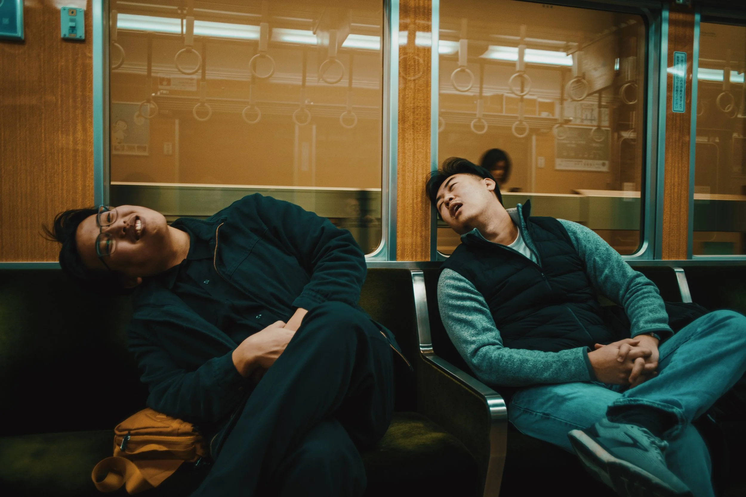 Two men sitting on a train, sleeping with heads leaning against a window, relaxed and resting.