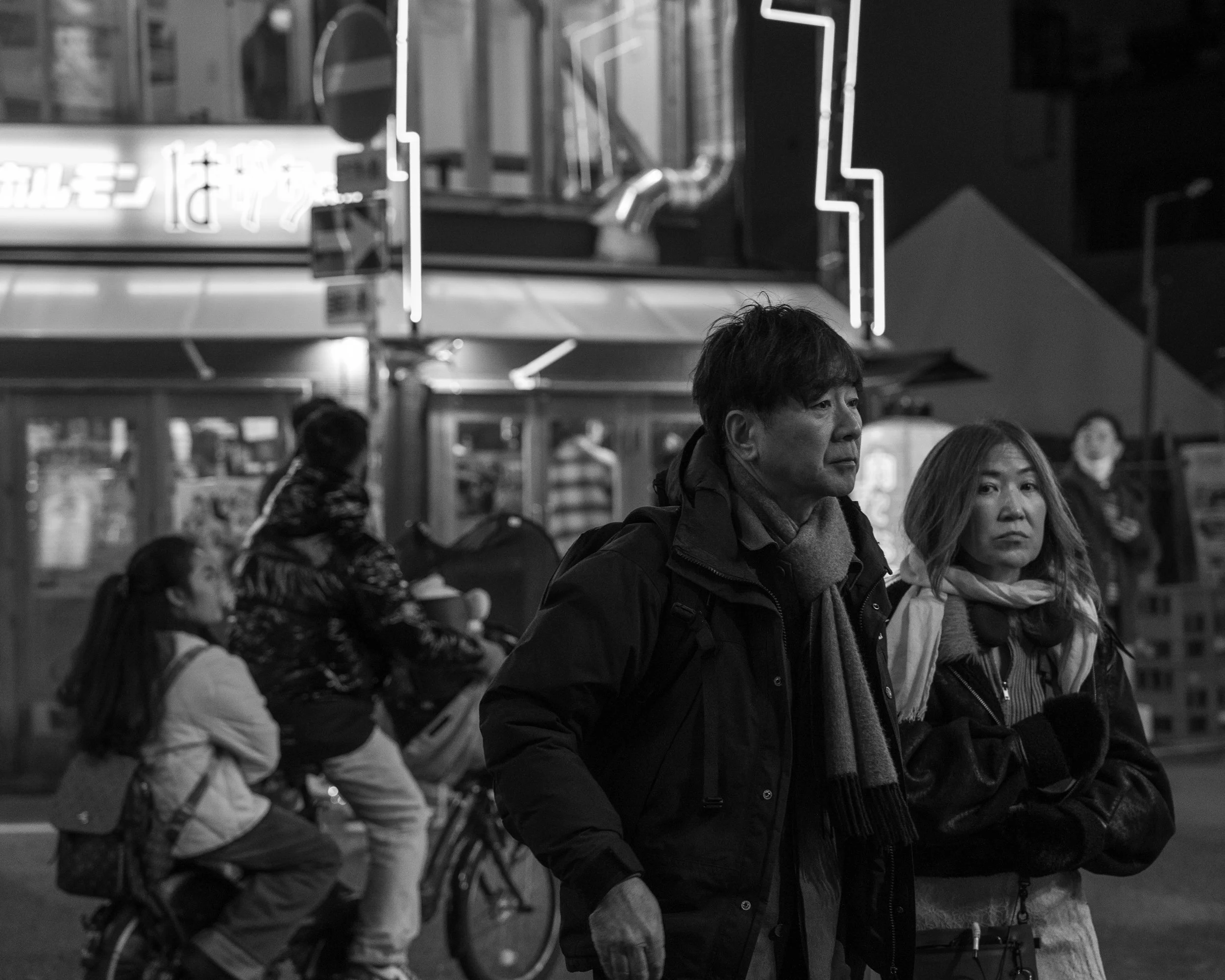 Black and white photo of a man and woman walking side by side on a city street at night. In the background, a woman and girl are riding a bicycle, and there are illuminated signs and buildings.