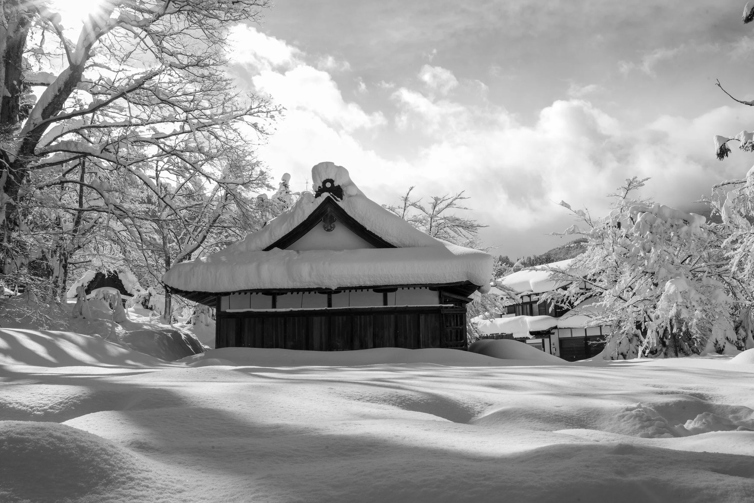 A snow-covered traditional Japanese building surrounded by snow-laden trees and a cloudy sky.