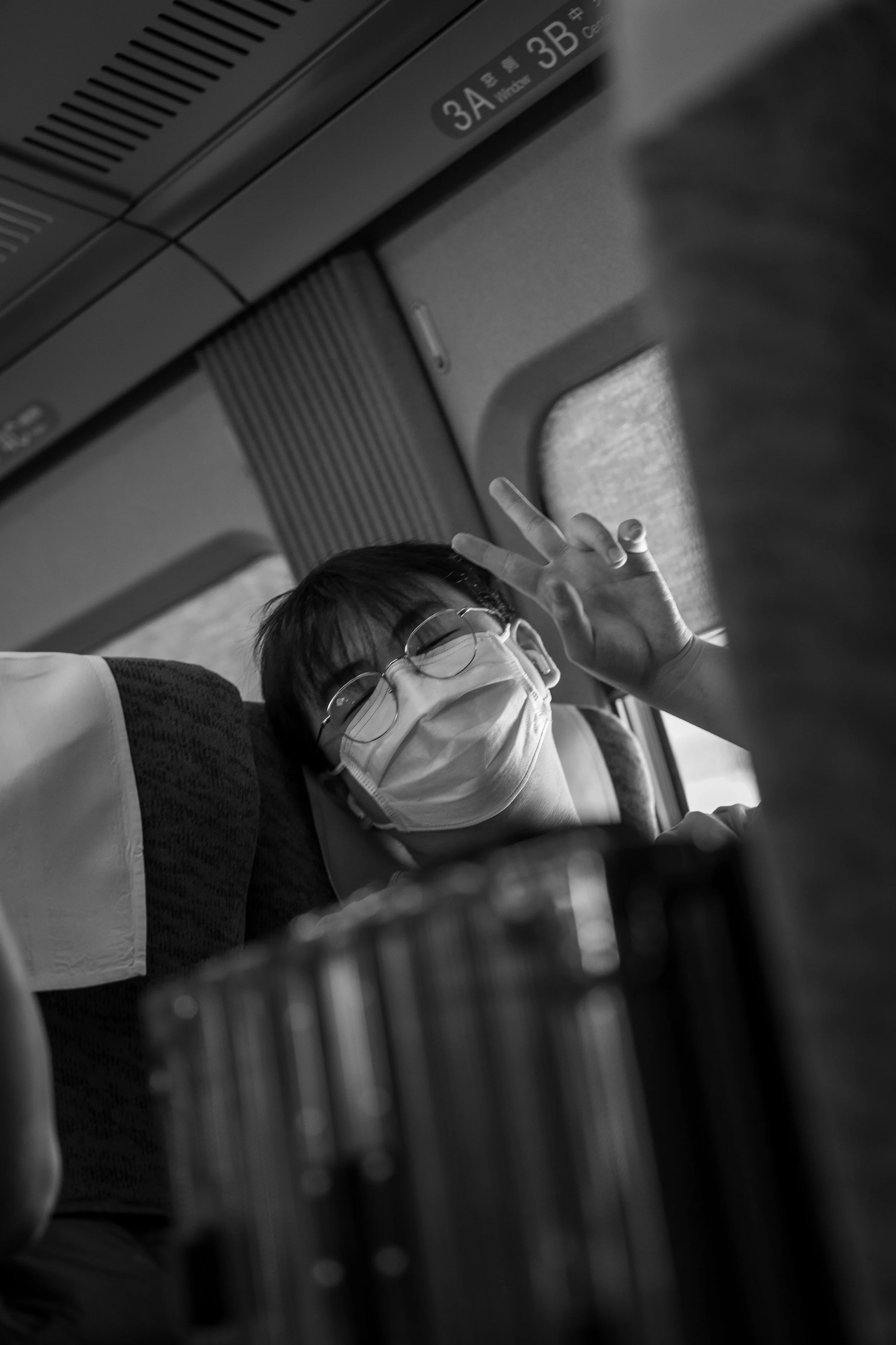 A woman wearing glasses and a face mask smiling and making a peace sign with her fingers while sitting on a bus.