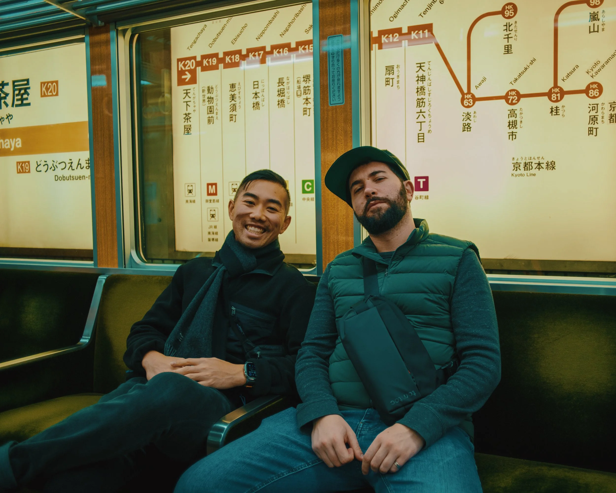 Two men sitting on a green bench inside a train station, with large Japanese and English train maps on the wall behind them.