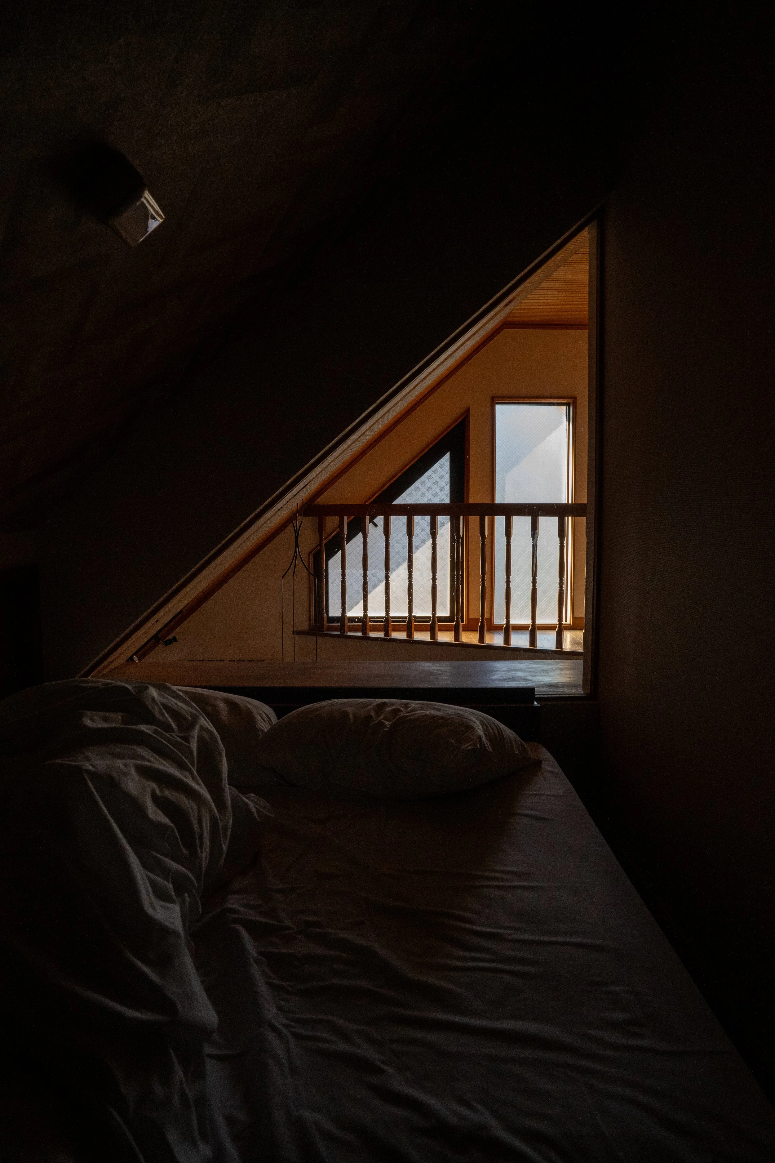 A dark bedroom with an unmade bed in the foreground and a small balcony with a wooden railing and frosted glass window in the background.