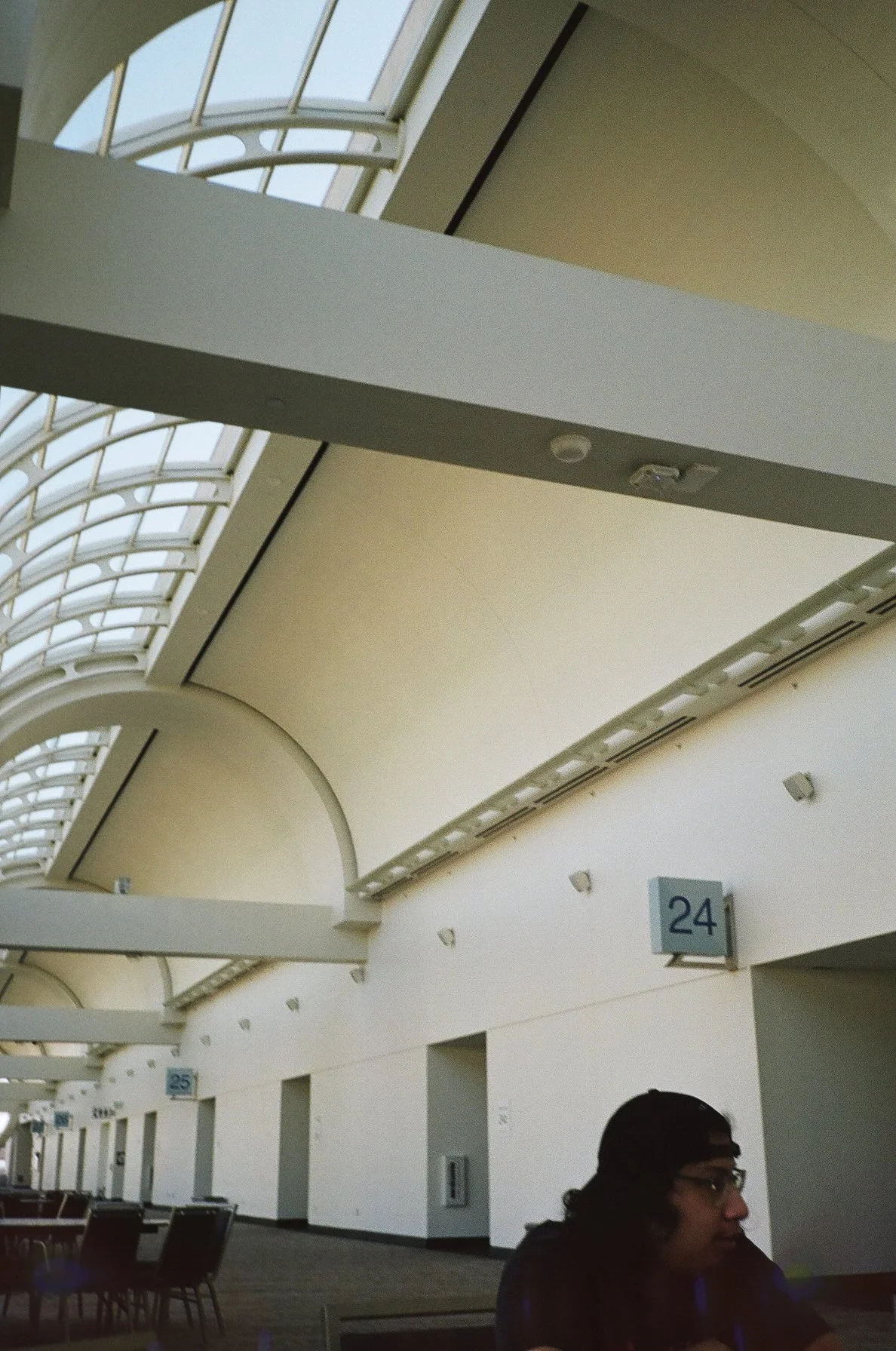 Interior of an airport or train station with high arched windows, numbered signs 24 and 25, and seating areas. A person wearing glasses and a cap is seated in the lower right corner.